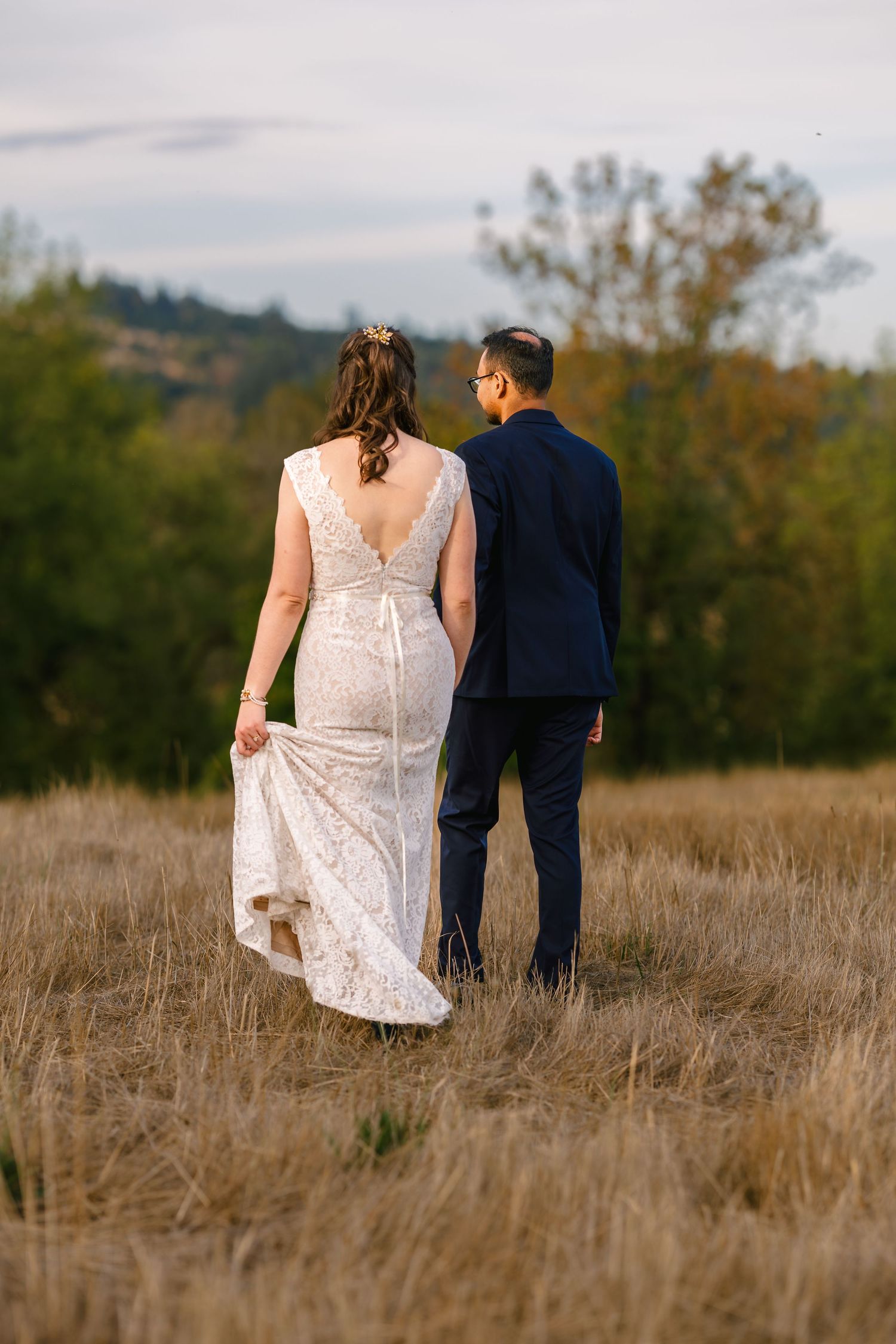 A couple walks through a golden field at sunset wearing formal wedding attire.