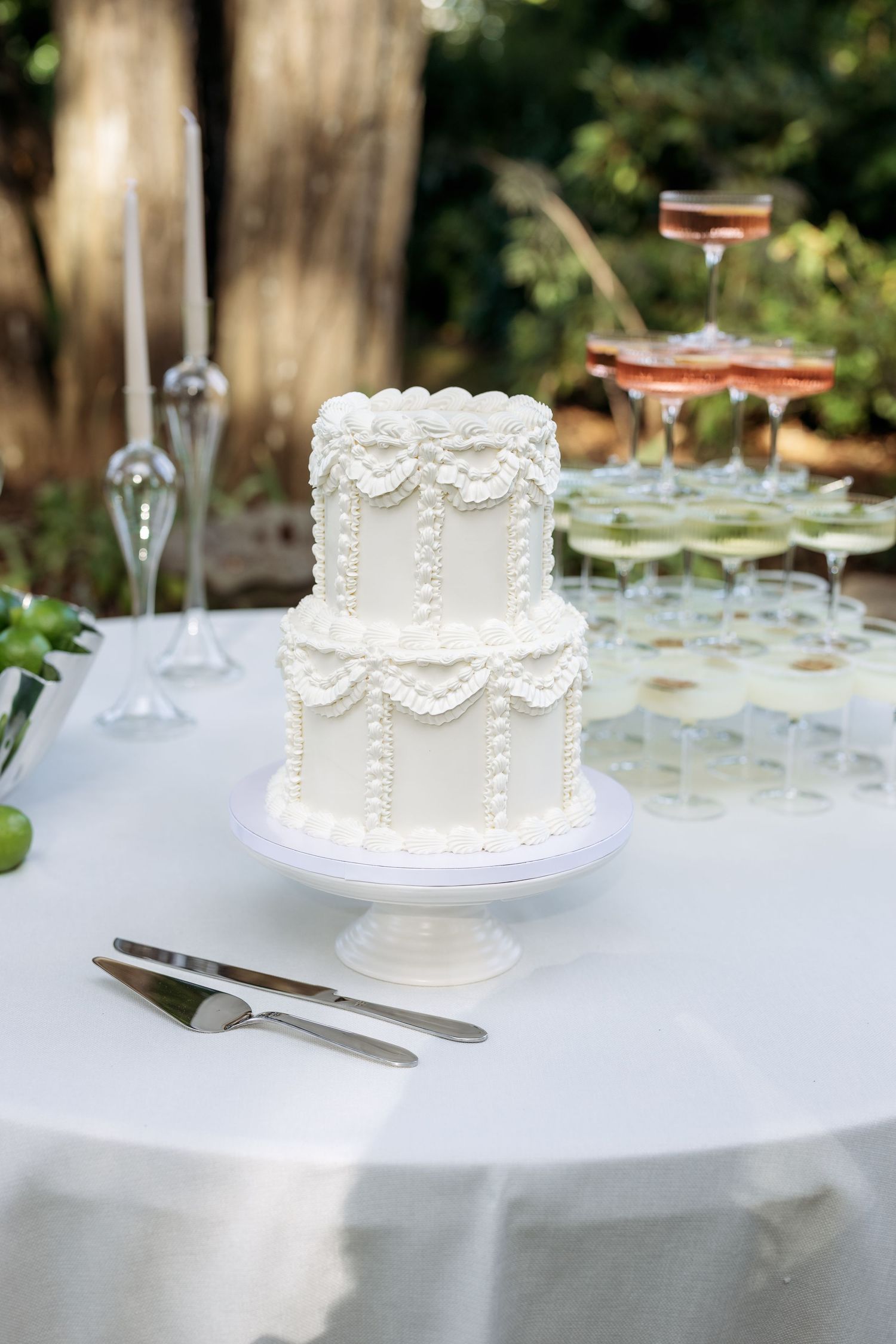 White tiered wedding cake with delicate lace piping and pearls displayed on an elegant outdoor table setting.
