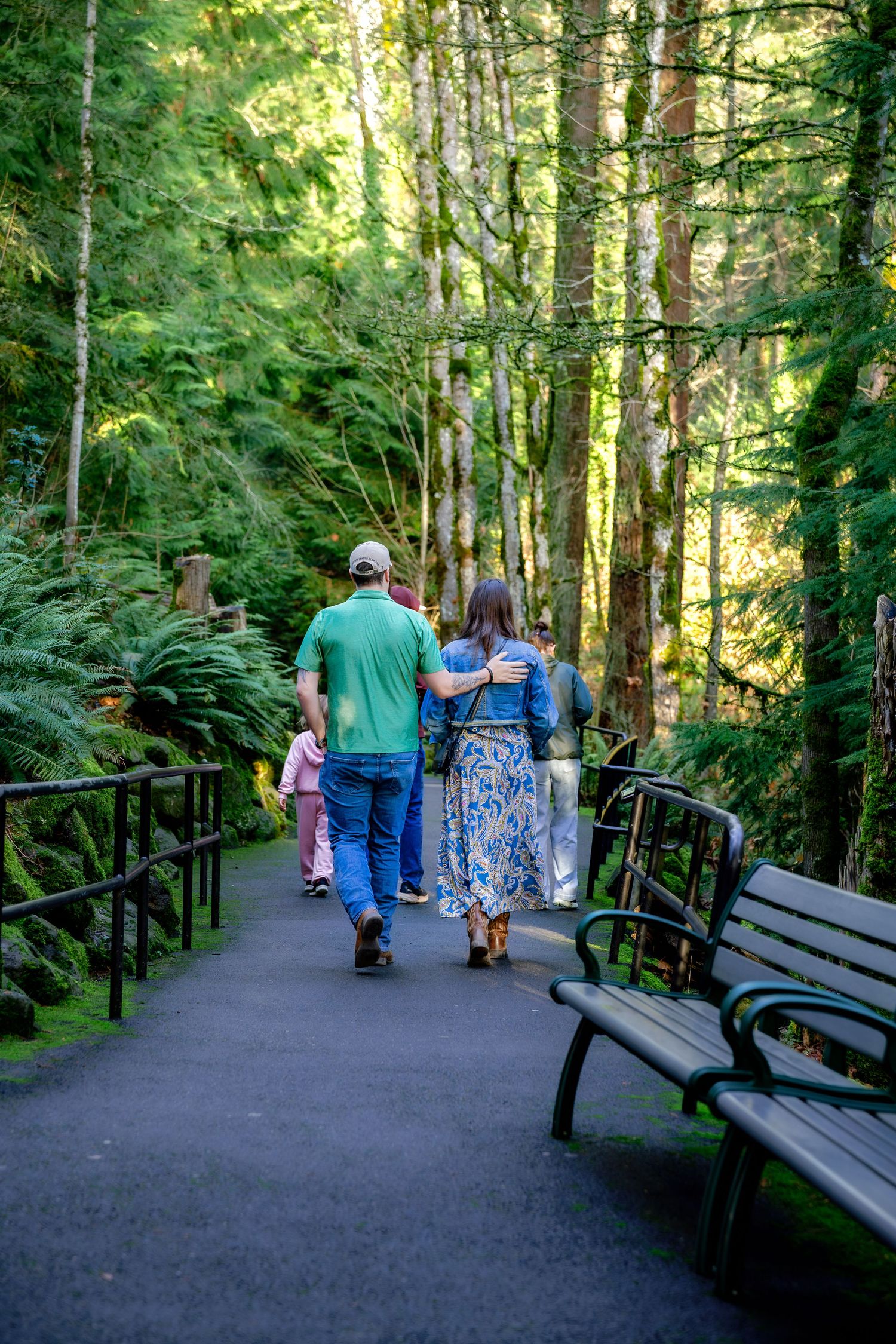 People walking along a shaded forest trail with wooden railings and benches in a lush green setting.