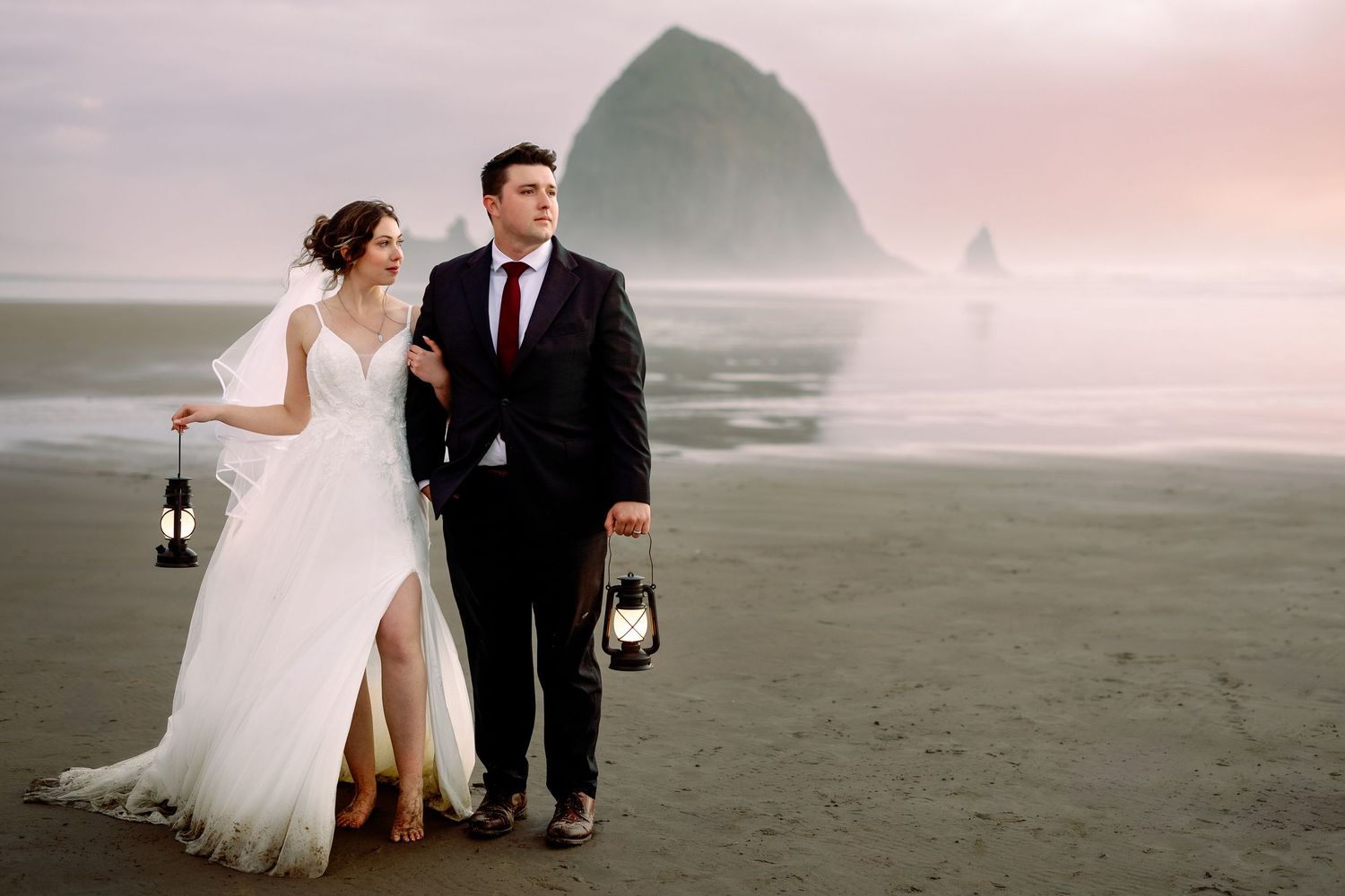 A couple in wedding attire walks together on a misty beach at sunset with dramatic cloudy skies.