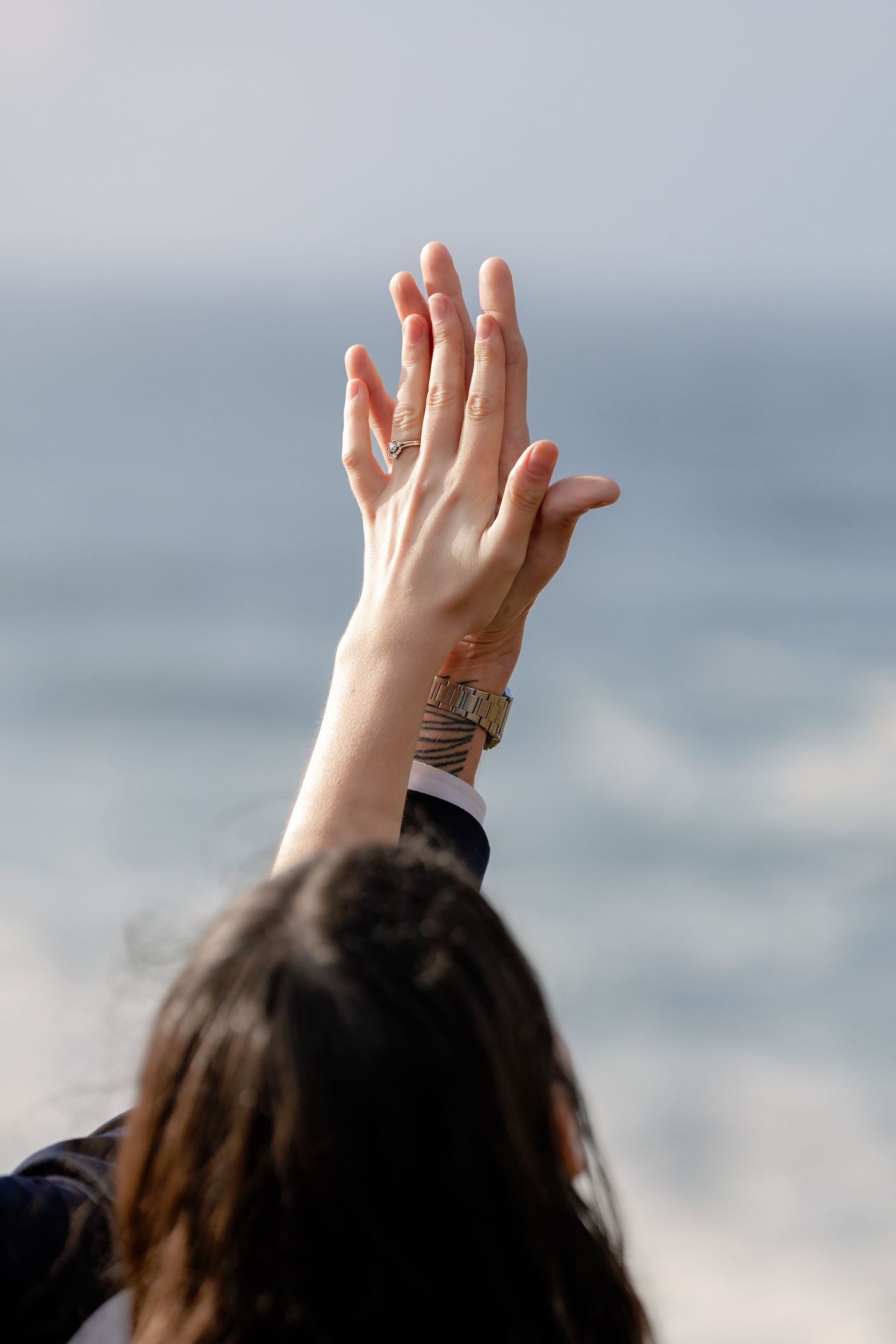 A raised hand with a wedding ring is silhouetted against a soft, cloudy sky.