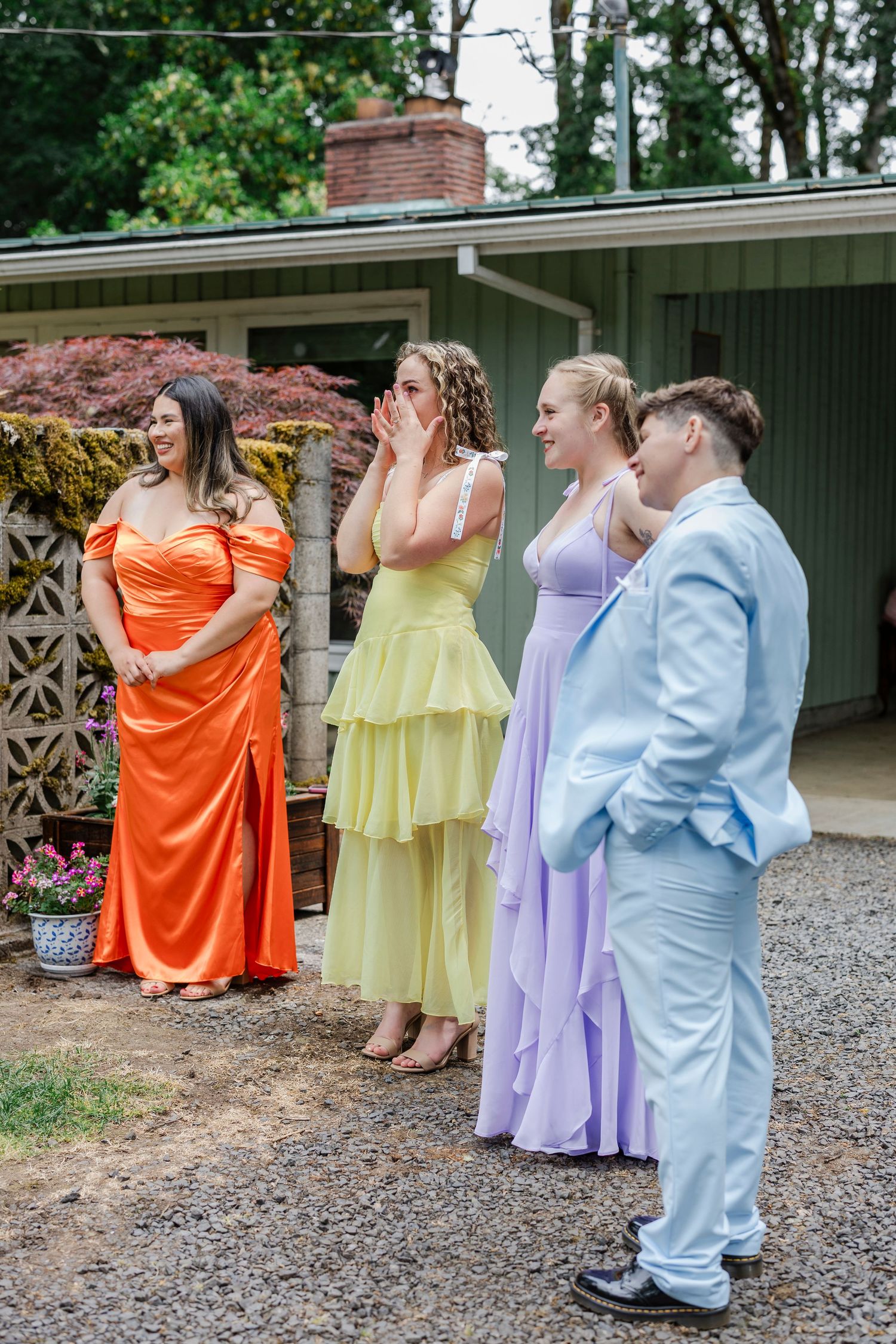 A group in colorful formal attire - orange, yellow, lavender and light blue - stand in a line on a gravel path.