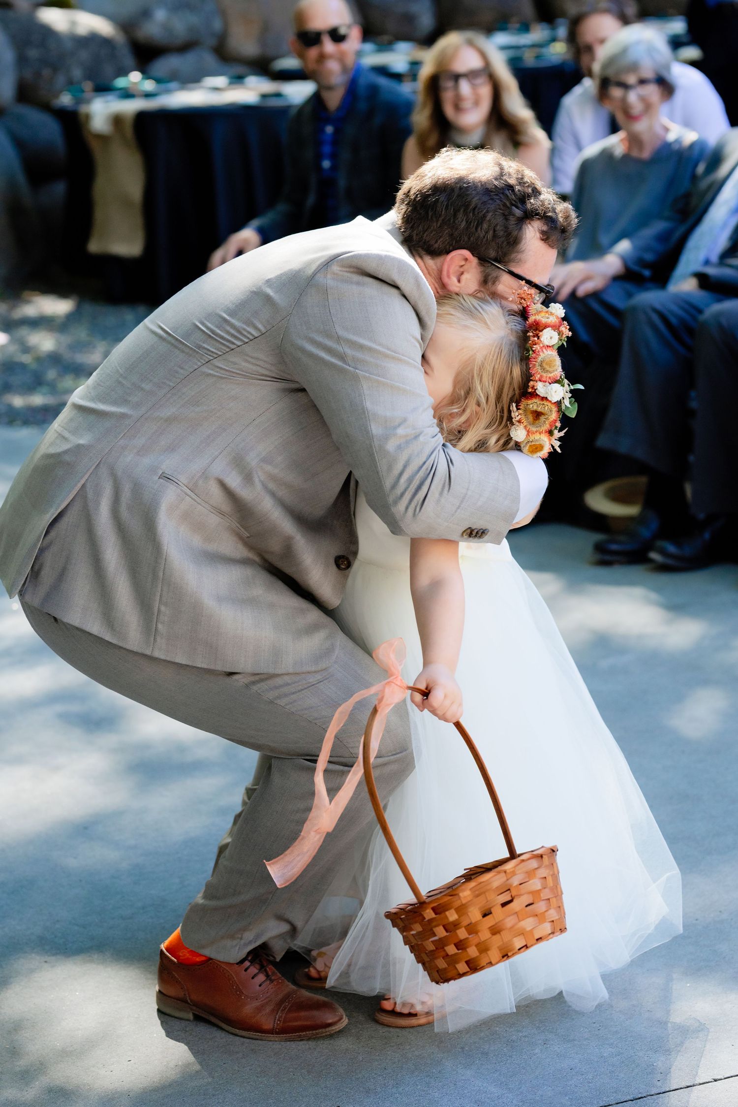 Groom in gray suit hugs young flower girl holding wicker basket during outdoor wedding ceremony.