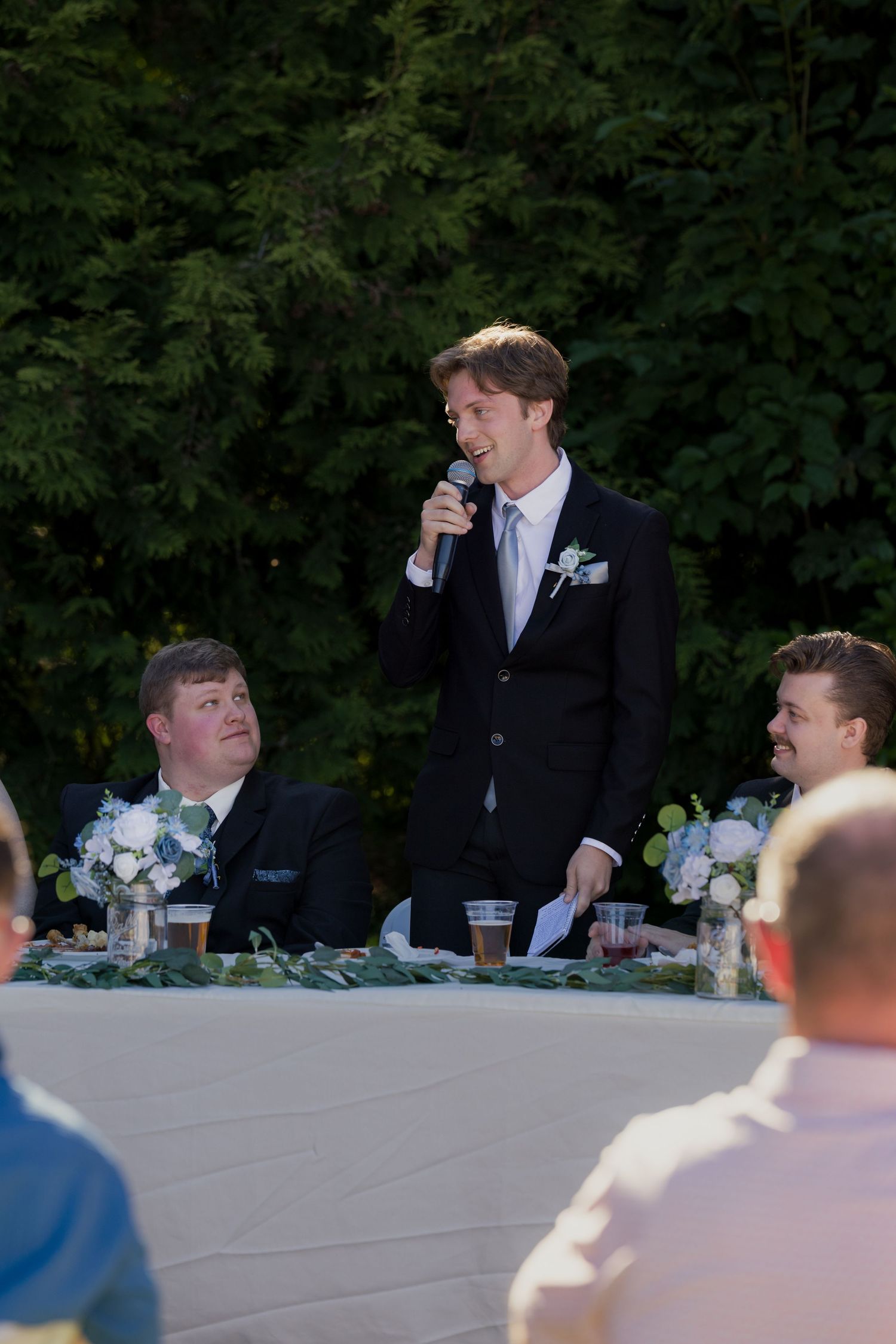 Smiling best man delivers a wedding speech into a microphone at an outdoor reception dinner.