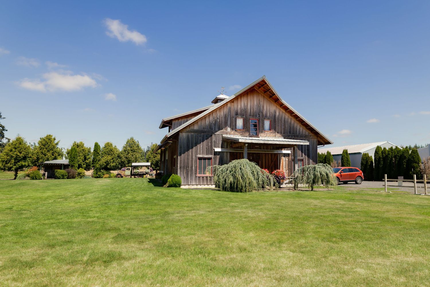 Rustic wooden barn-style home with a large green lawn, surrounded by trees under a bright blue sky.