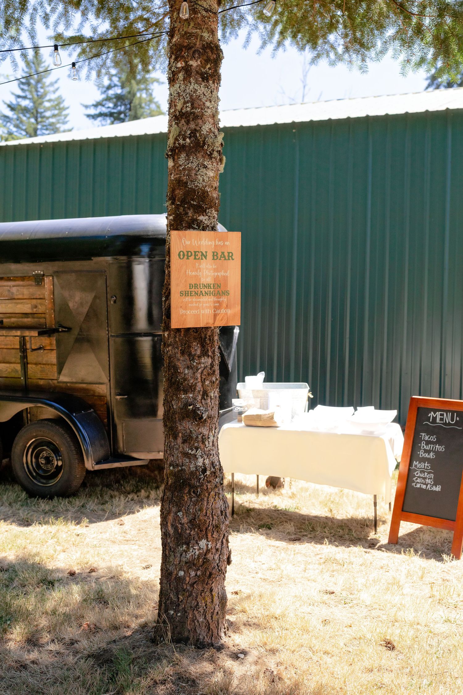 Open bar sign on a tree at an outdoor wedding venue with a mobile bar and menu chalkboard.