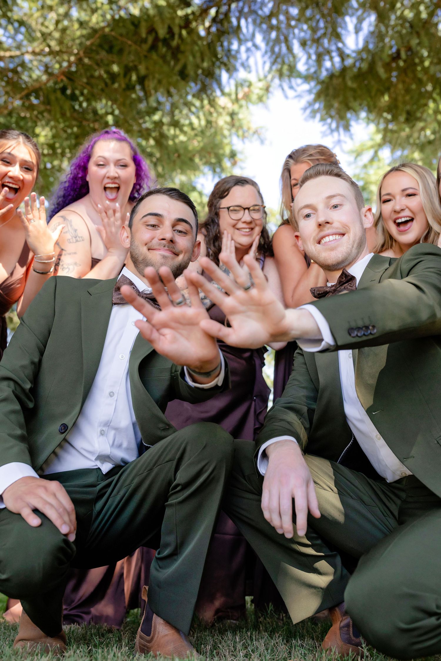 Two grooms show off wedding rings surrounded by cheering wedding party members under green trees.