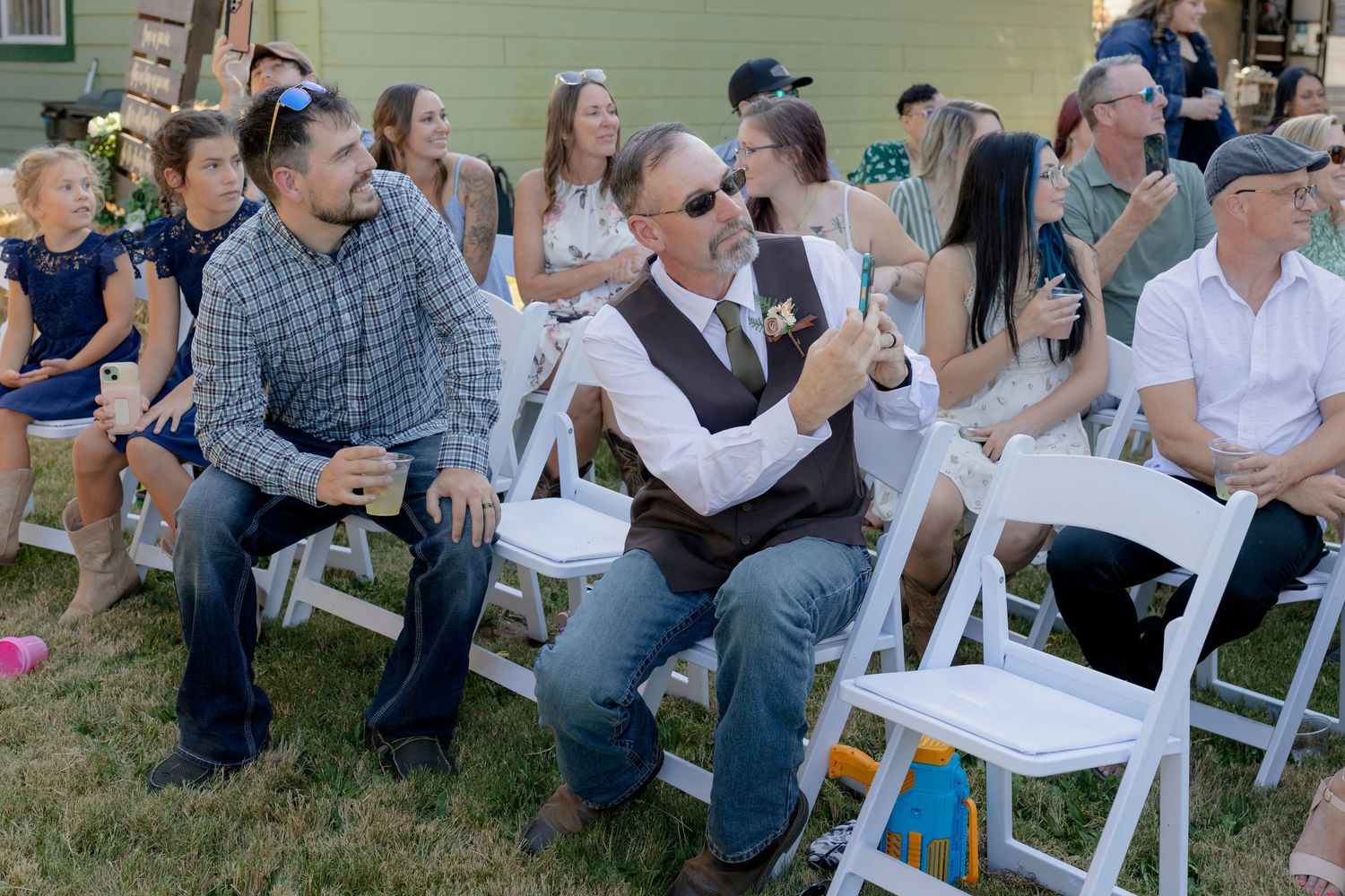 Wedding guests including two men in vests seated in white chairs at an outdoor ceremony.