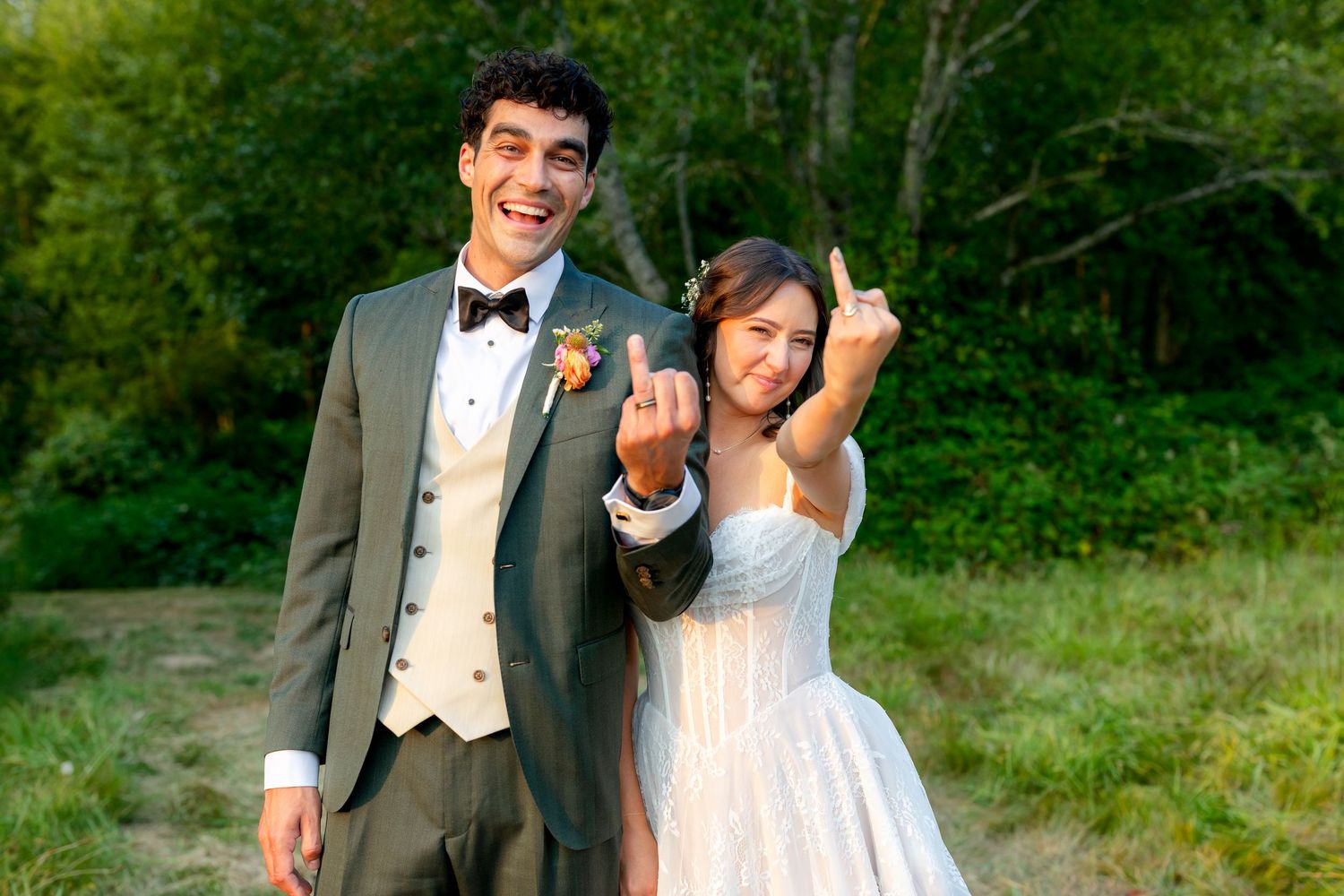 Newlyweds laugh and gesture playfully outdoors in wedding attire, groom in gray suit and bride in white dress.