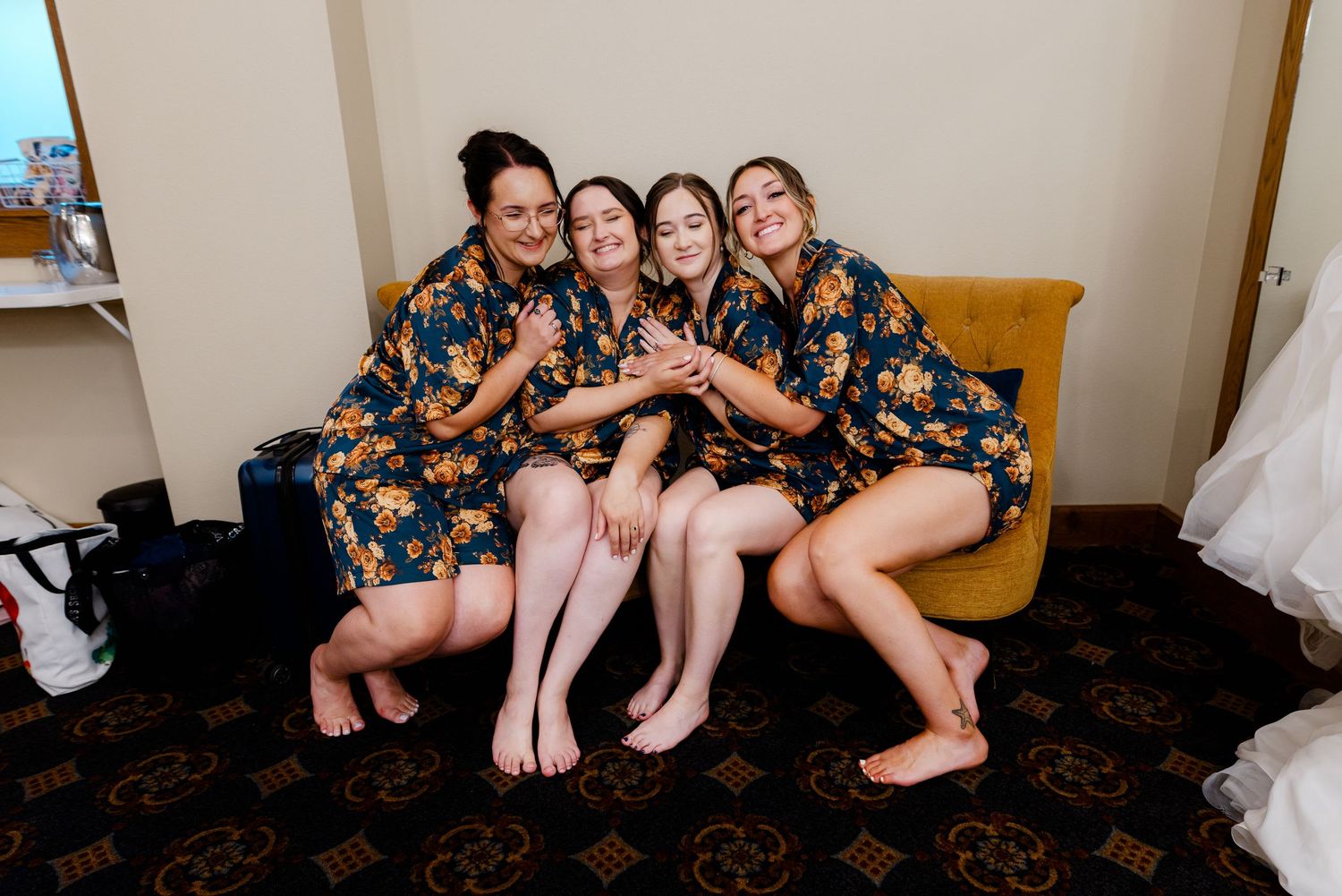 Four bridesmaids in matching floral robes sitting together smiling and hugging on a couch before the wedding.