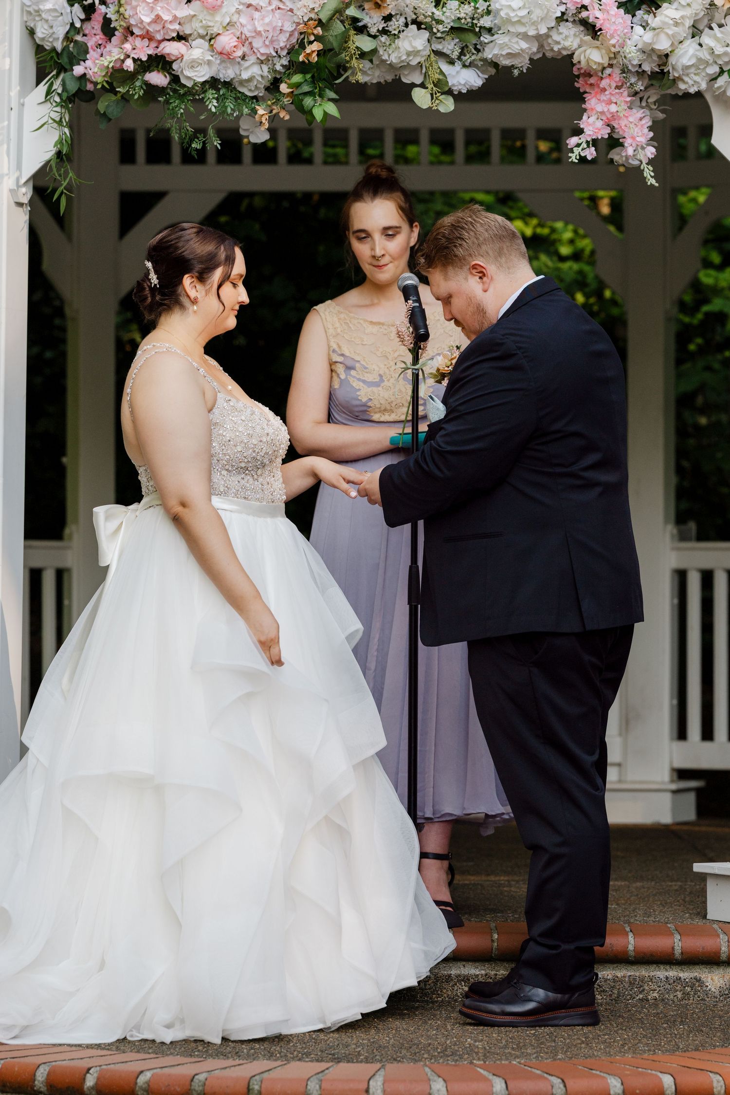 Bride and groom exchange rings at outdoor gazebo wedding ceremony officiated by woman in lavender dress.