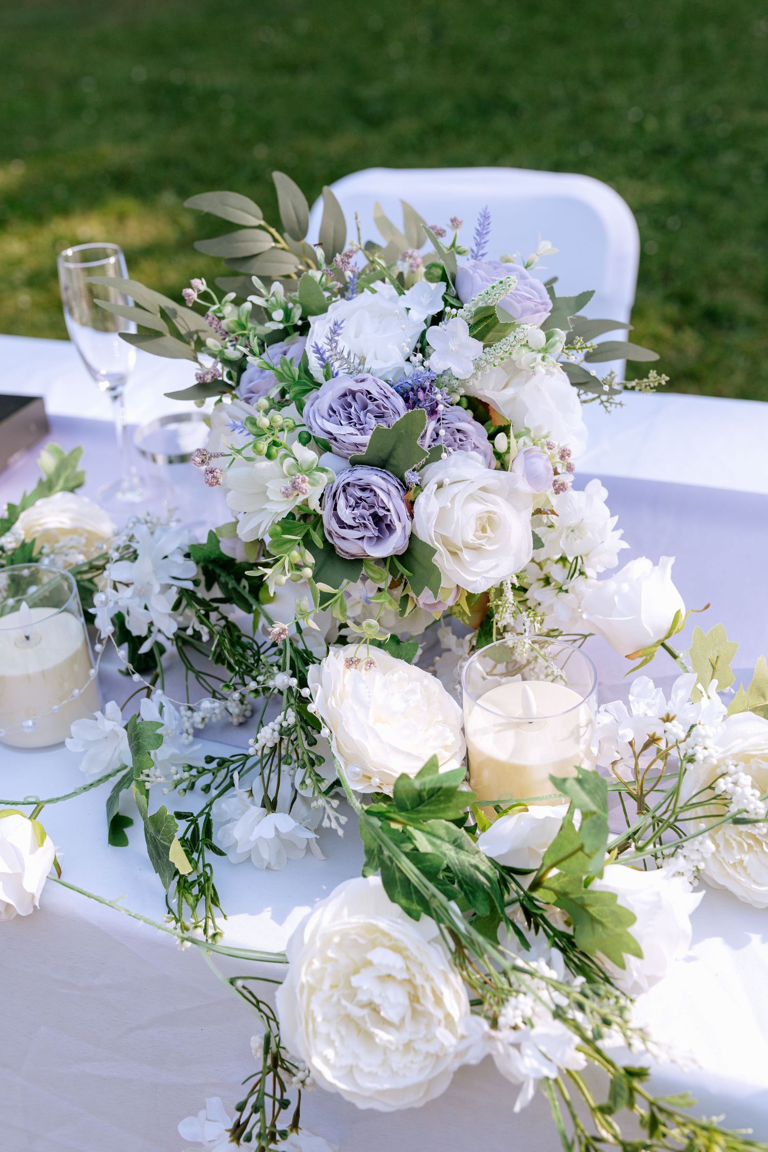 Elegant outdoor wedding table centerpiece featuring white and lavender roses with candles and greenery.