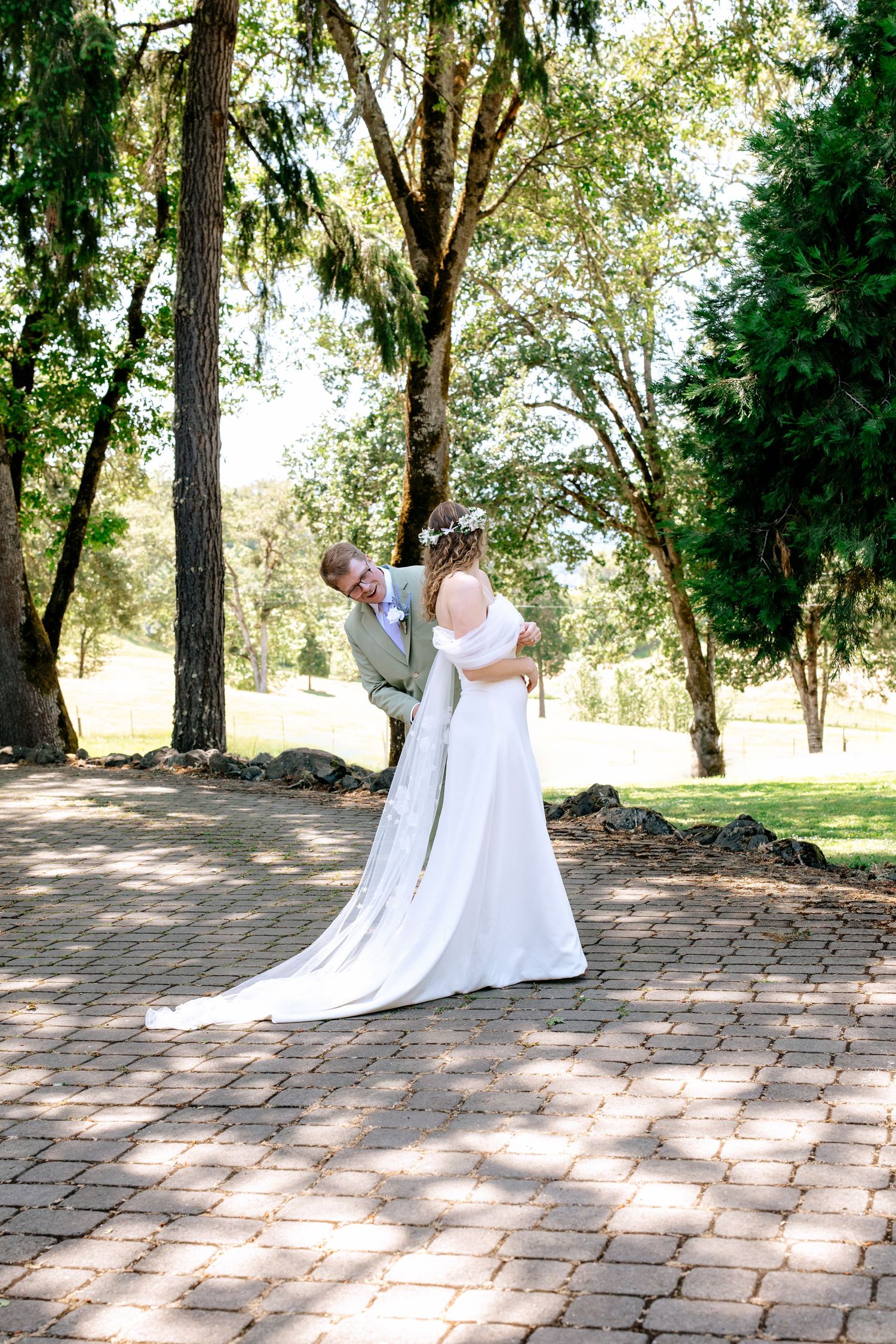 Bride and groom share a romantic embrace on a brick pathway surrounded by lush green trees at an outdoor wedding.