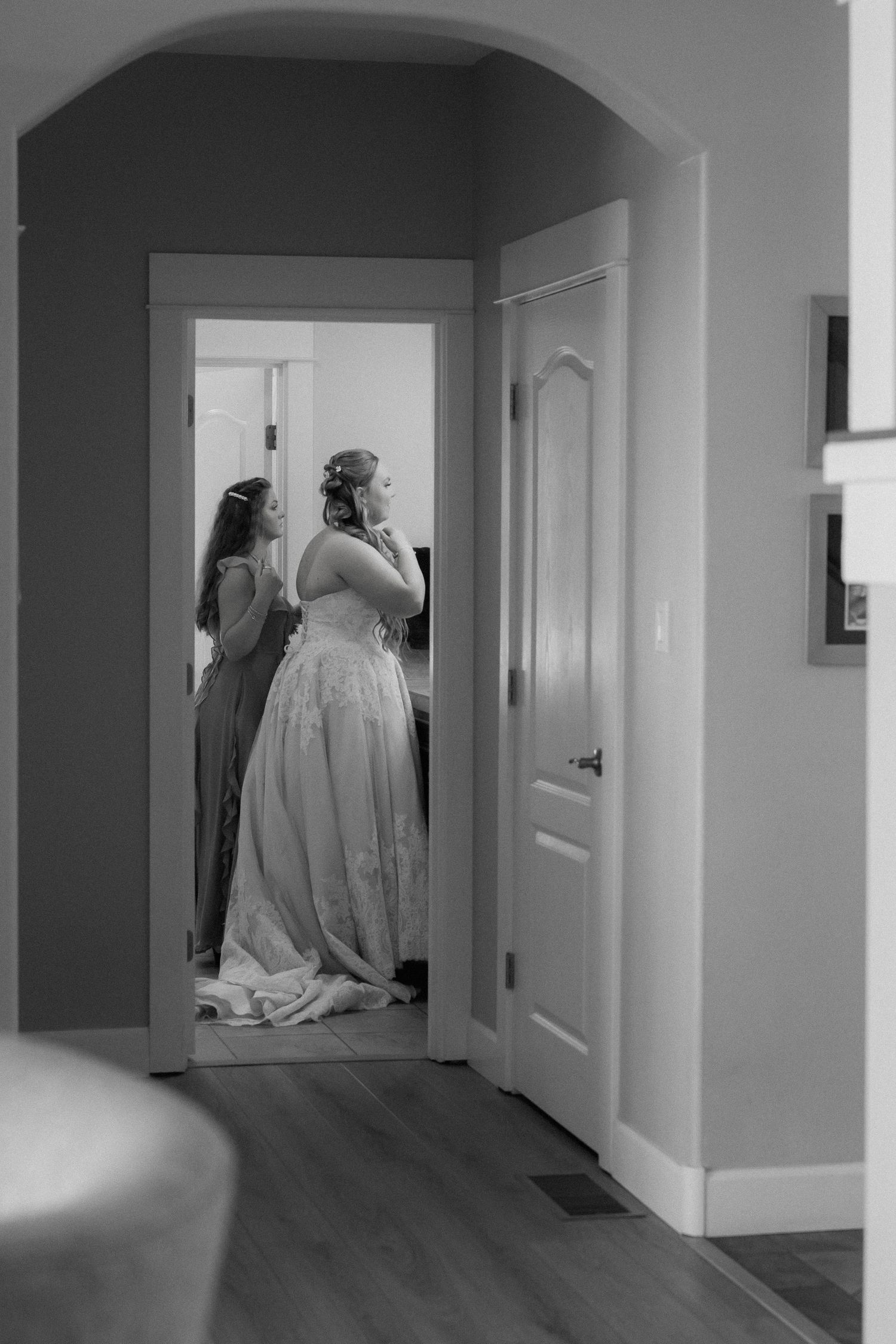 Black and white photo of bride in lace gown being helped by bridesmaid while getting ready for wedding.