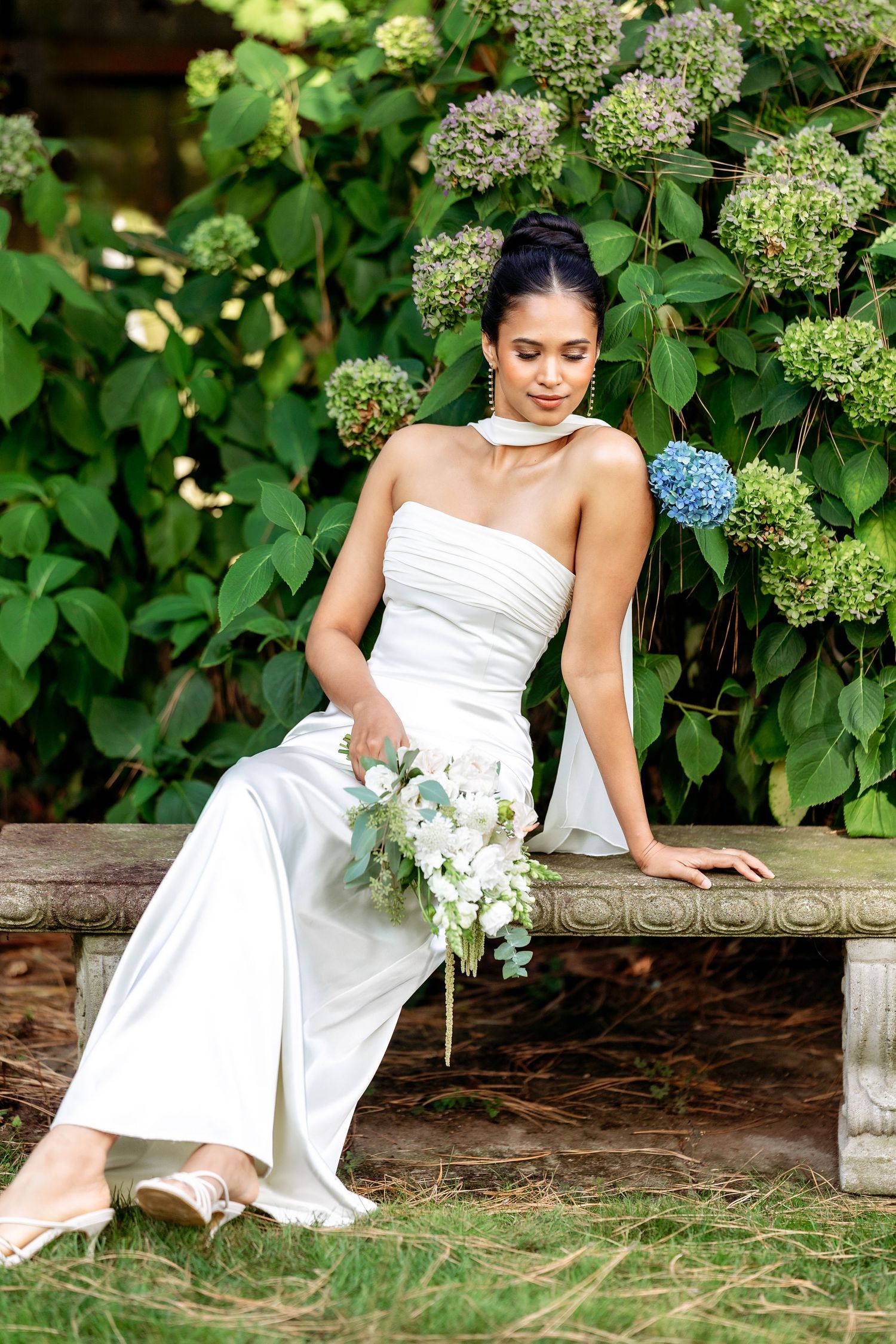 A person in a white wedding dress poses gracefully near blooming hydrangea bushes in a garden setting.