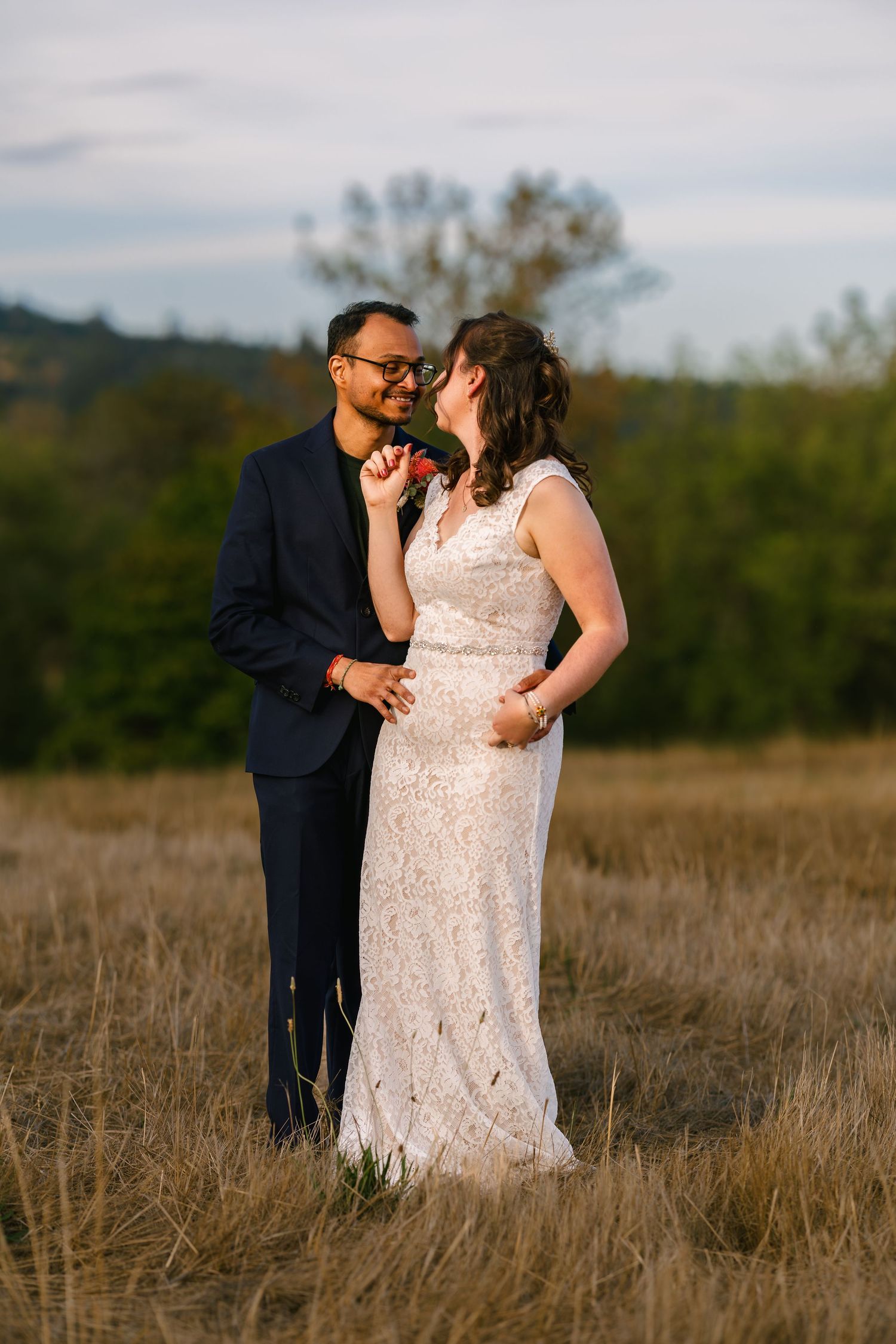 A romantic couple embraces in a field during golden hour wearing formal wedding attire.