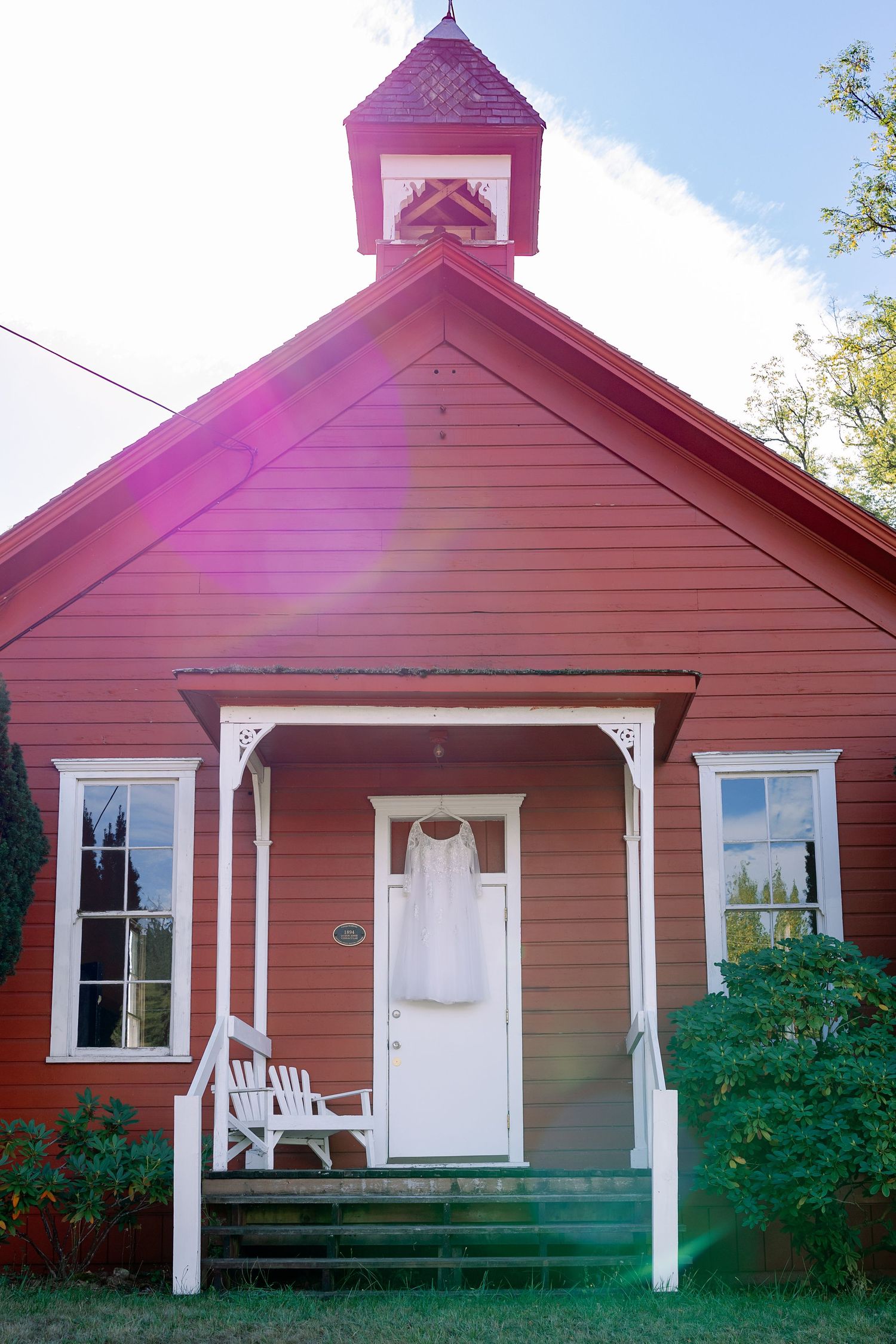 Red schoolhouse with white trim and pink steeple displaying a wedding dress on the front door.