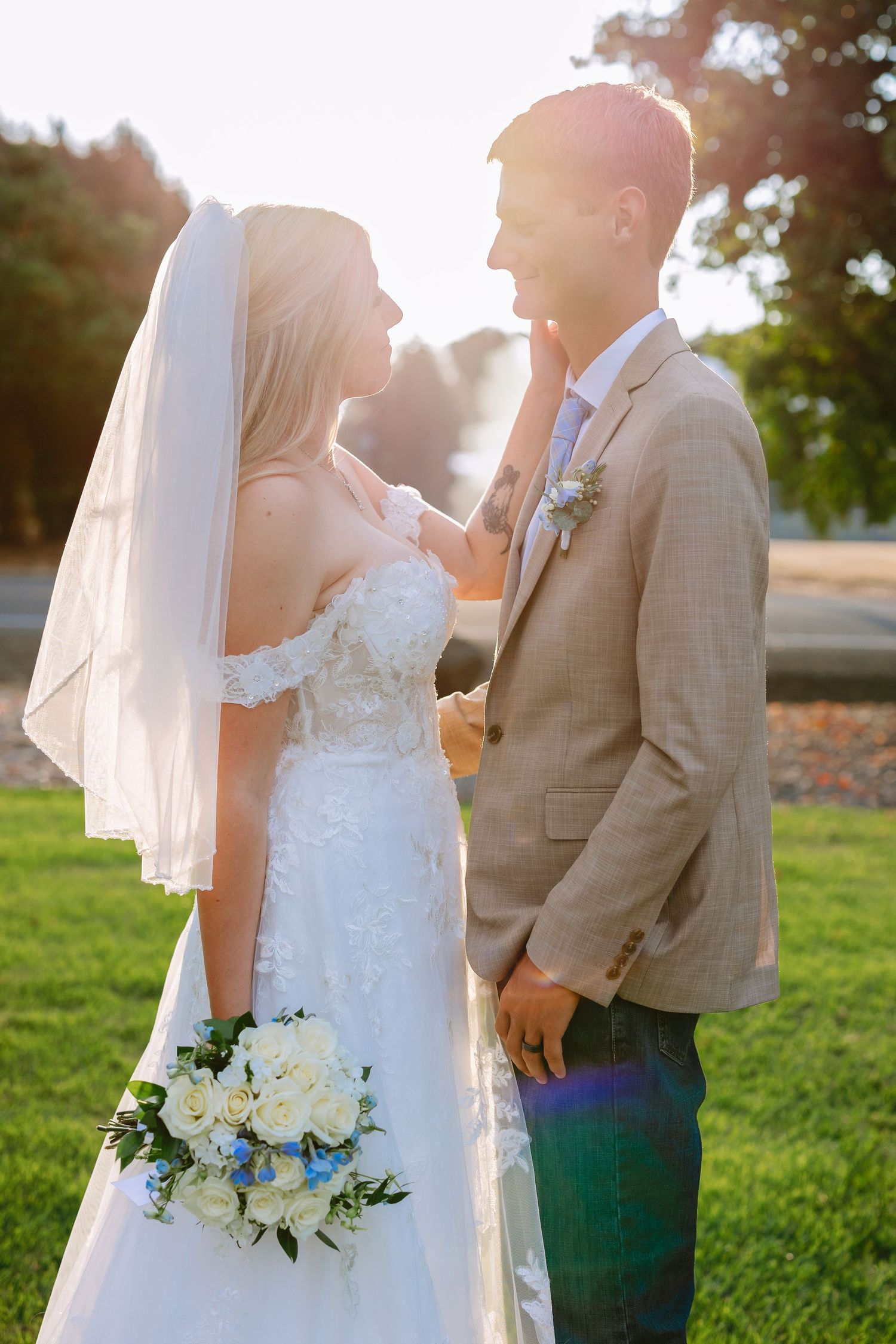 A bride in white dress holds a bouquet while sharing an intimate moment during sunset at an outdoor wedding.