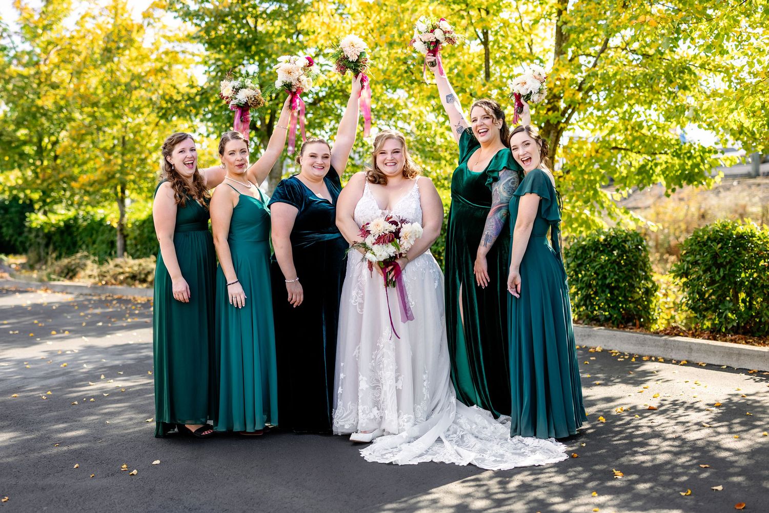 A bridal party in emerald green dresses and white wedding gown pose joyfully on a sunny outdoor path.