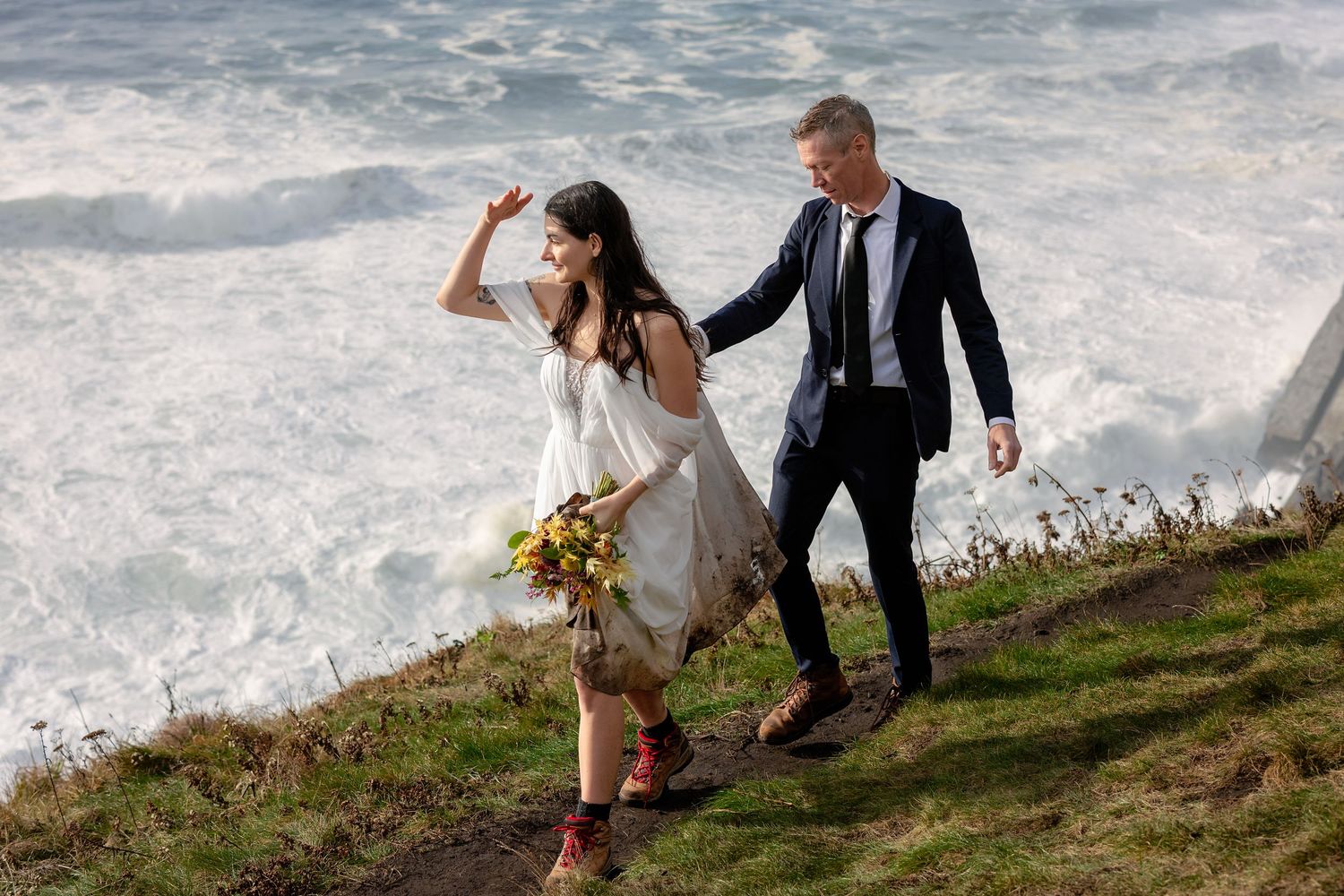 A couple in wedding attire walks along a scenic coastal cliff with crashing waves below.