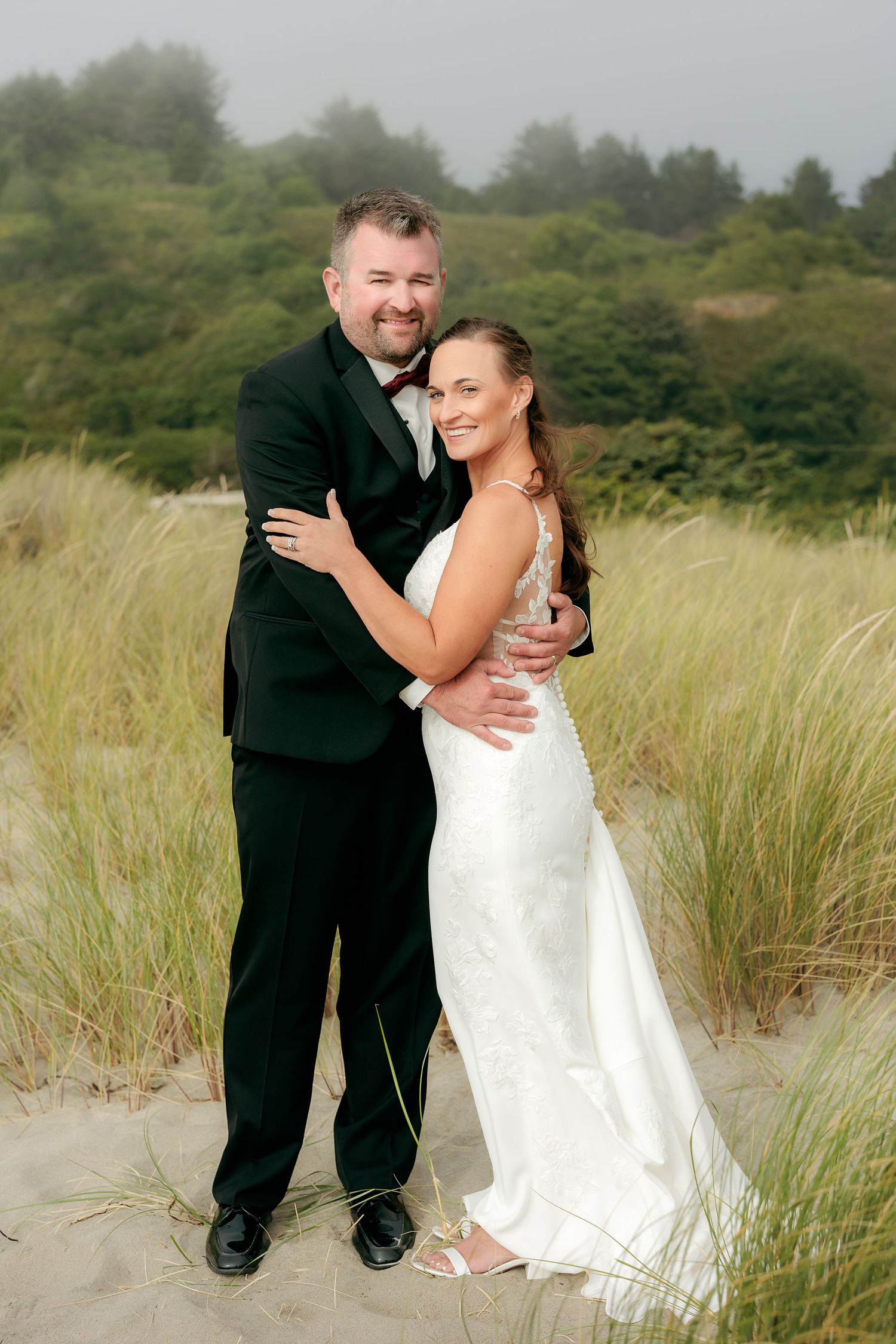Newlyweds embrace on beach with misty green hills in background.