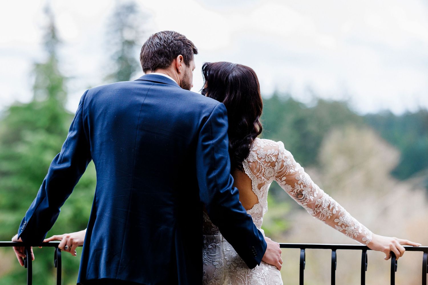 A newlywed couple in formal attire embrace while overlooking a scenic mountain view from a balcony railing.