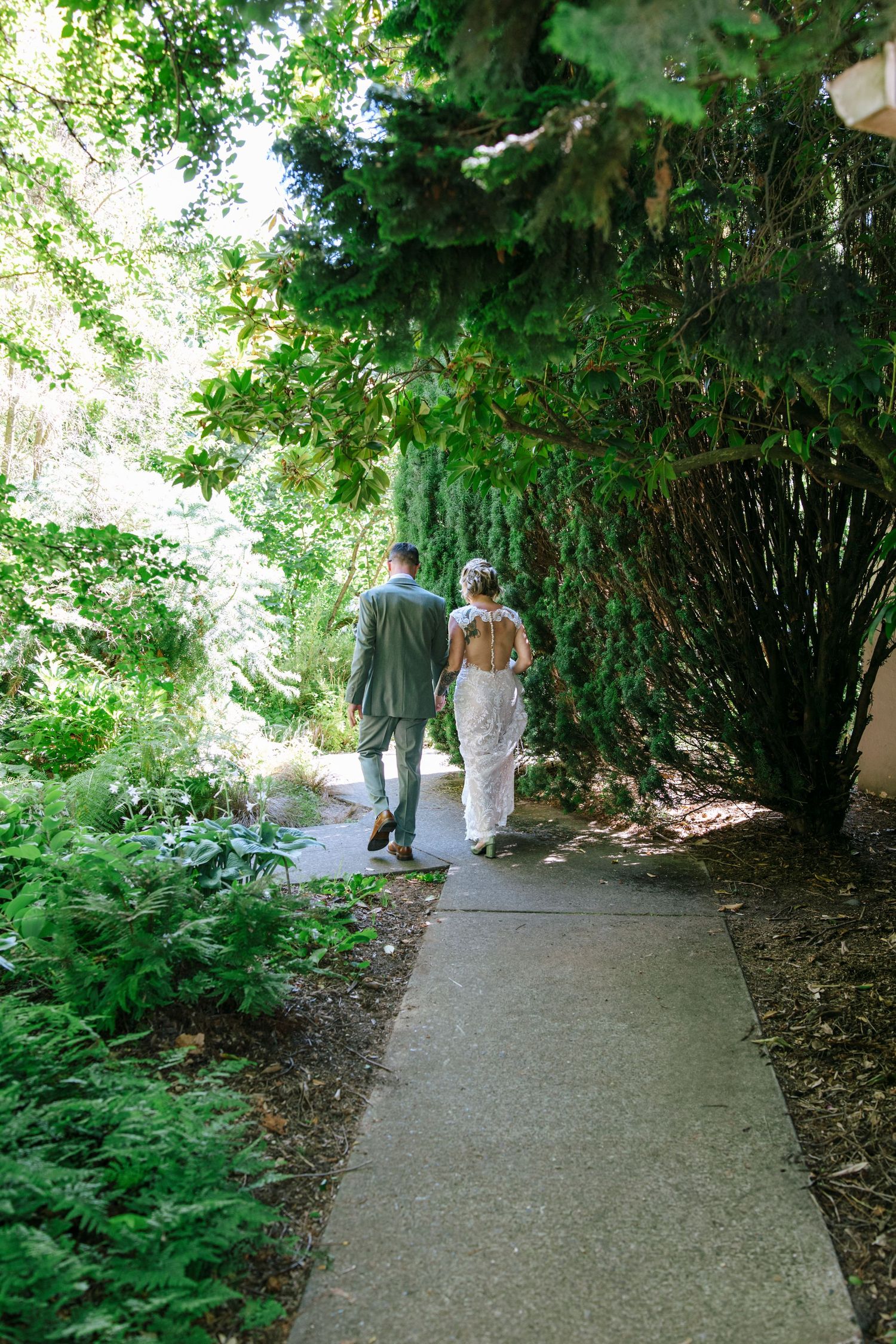 Wedding couple walking along a garden pathway surrounded by lush green vegetation.