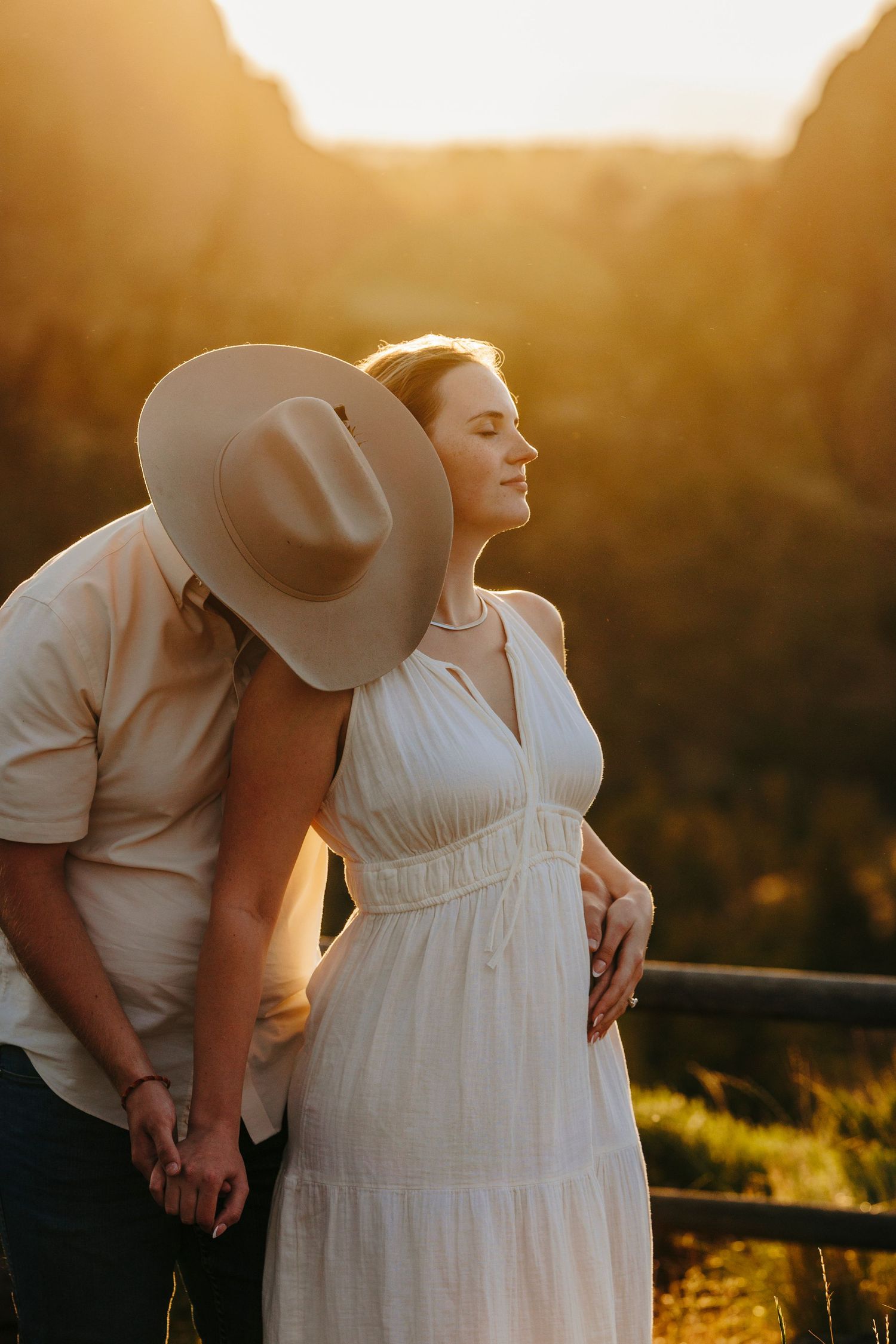 A couple in western wear embraces during golden hour with scenic mountain backdrop.