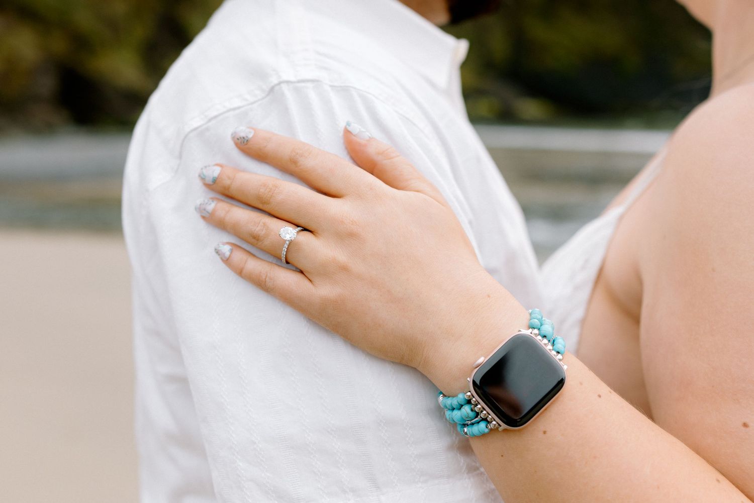 Woman's hand with ring and turquoise smartwatch band resting on man's white shirt at beach.