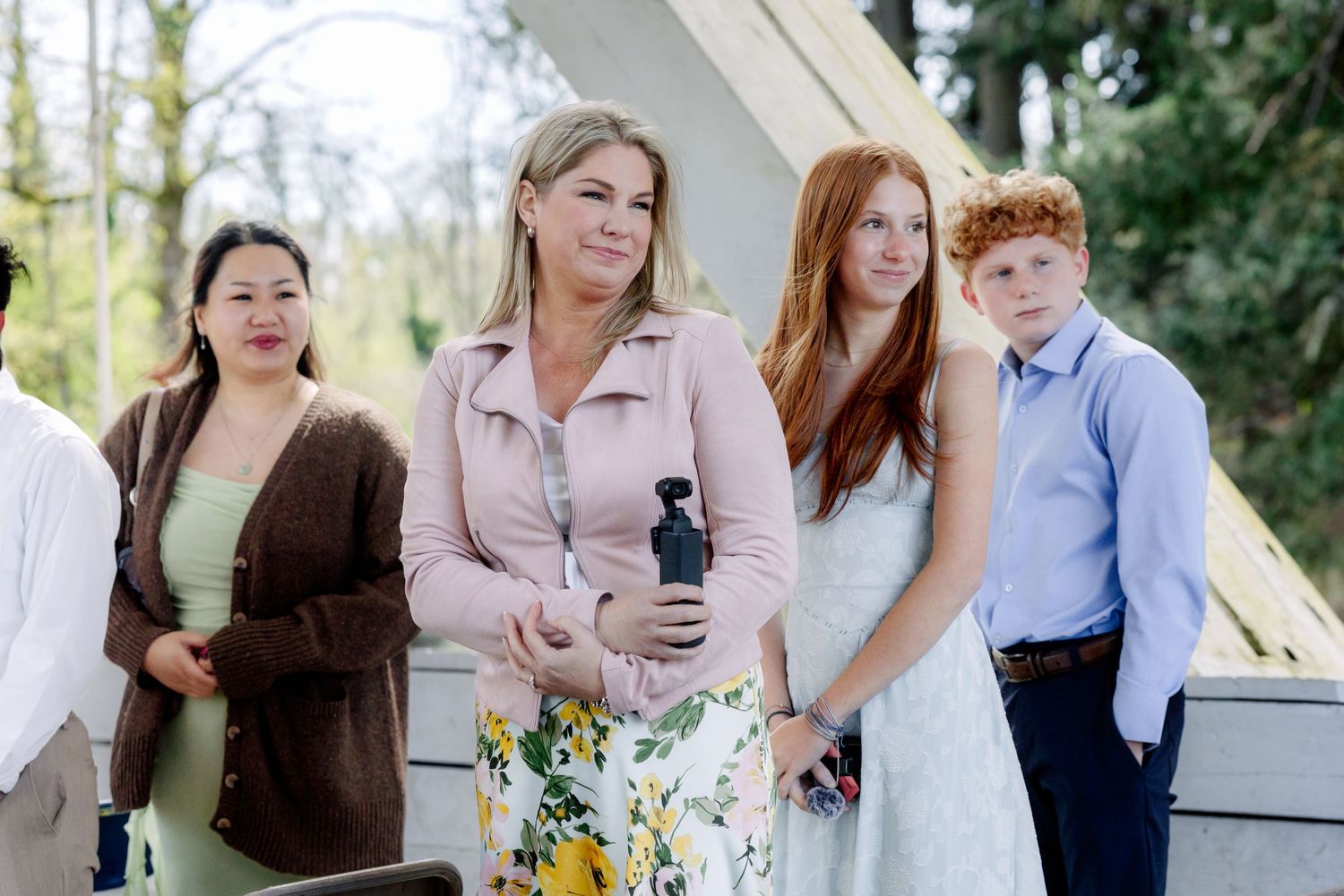 A group of four people including a woman in a pink jacket and floral dress standing outdoors at a wedding event.