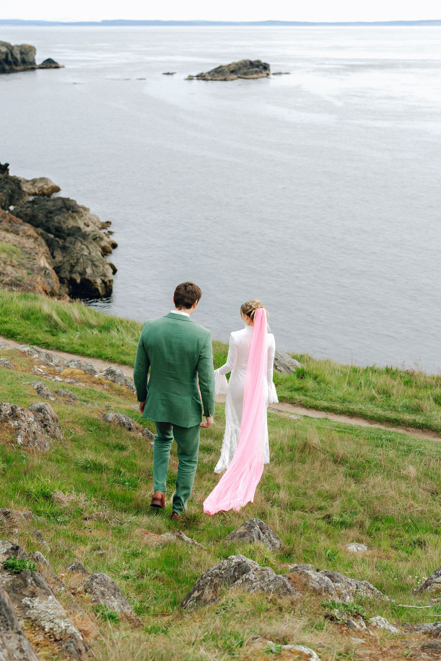 Bride in white dress with pink veil and groom in green suit walking hand in hand toward rocky ocean coastline.
