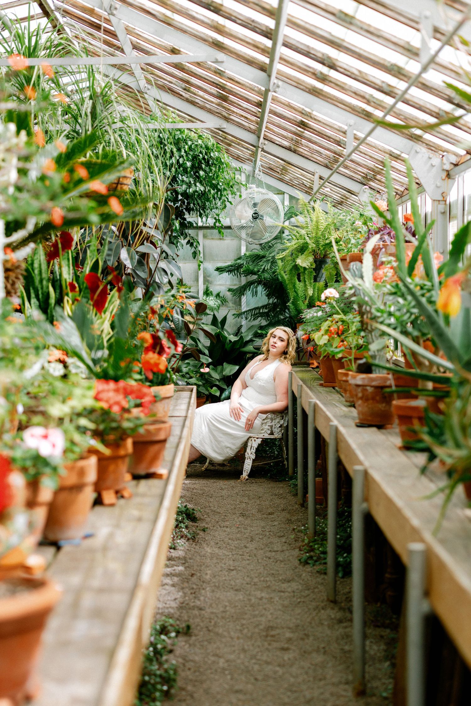 Blonde woman in white dress sitting among potted plants in a lush greenhouse aisle.
