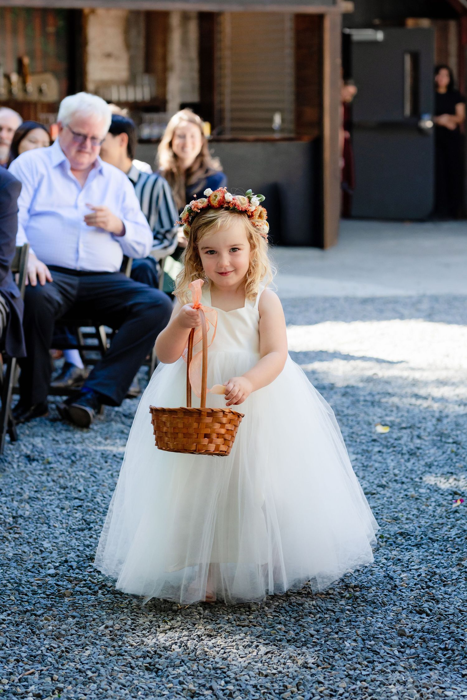 Flower girl in white tulle dress with floral crown carrying basket at wedding.