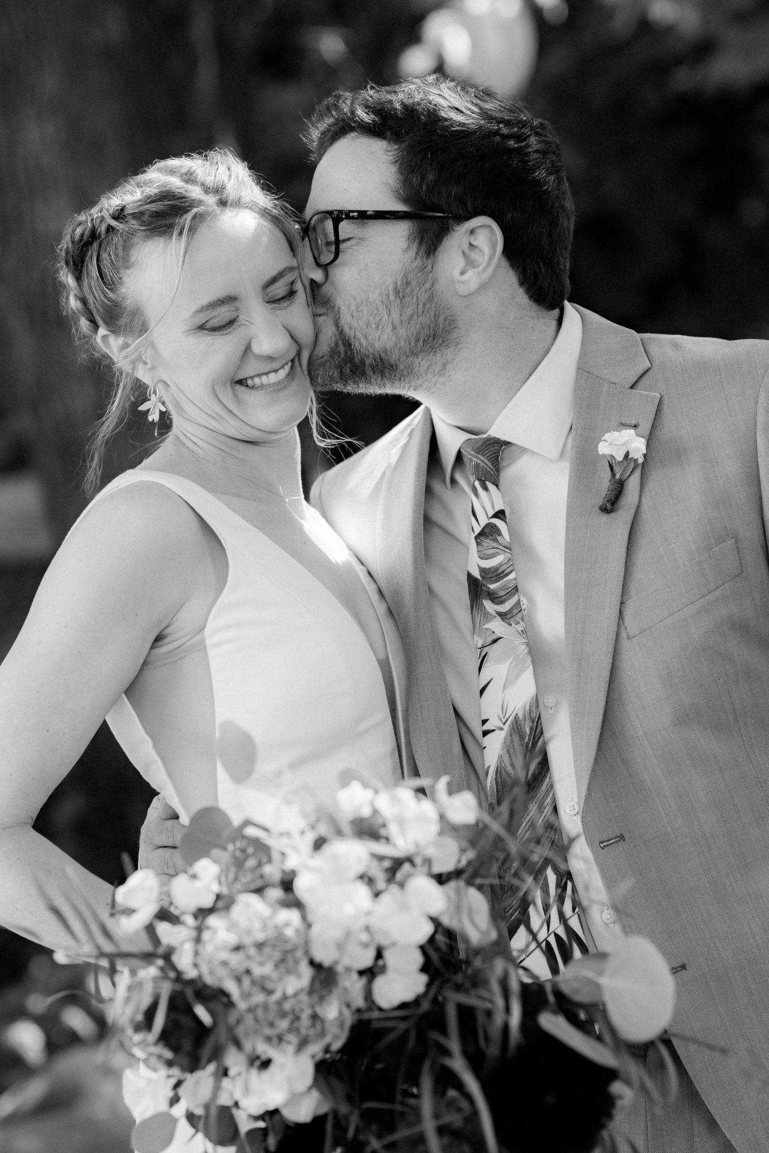 Groom in gray suit kisses bride on cheek as she smiles holding a floral bouquet in black and white wedding photo.