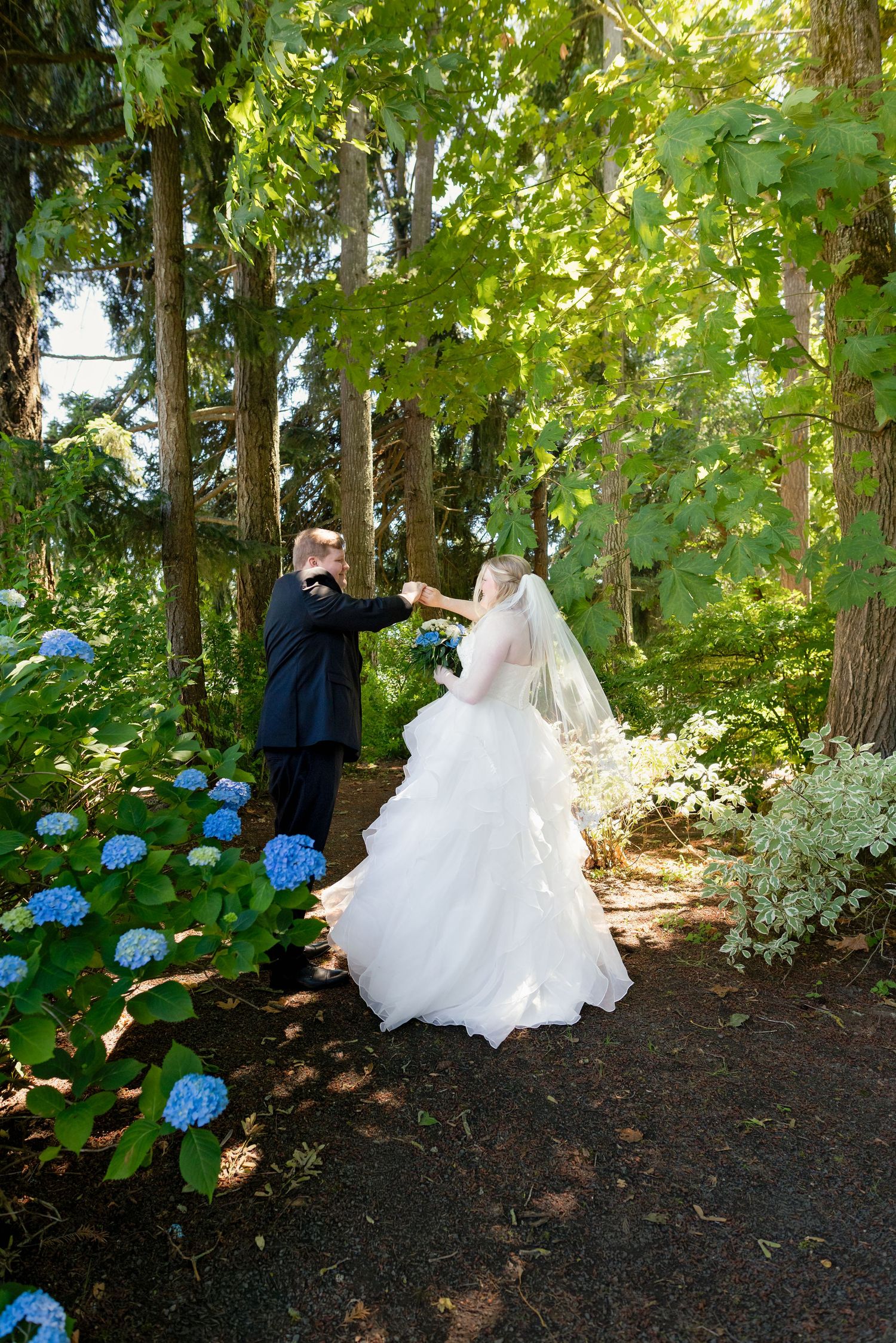 Bride and groom share a romantic moment dancing among blue hydrangeas in a lush garden setting.