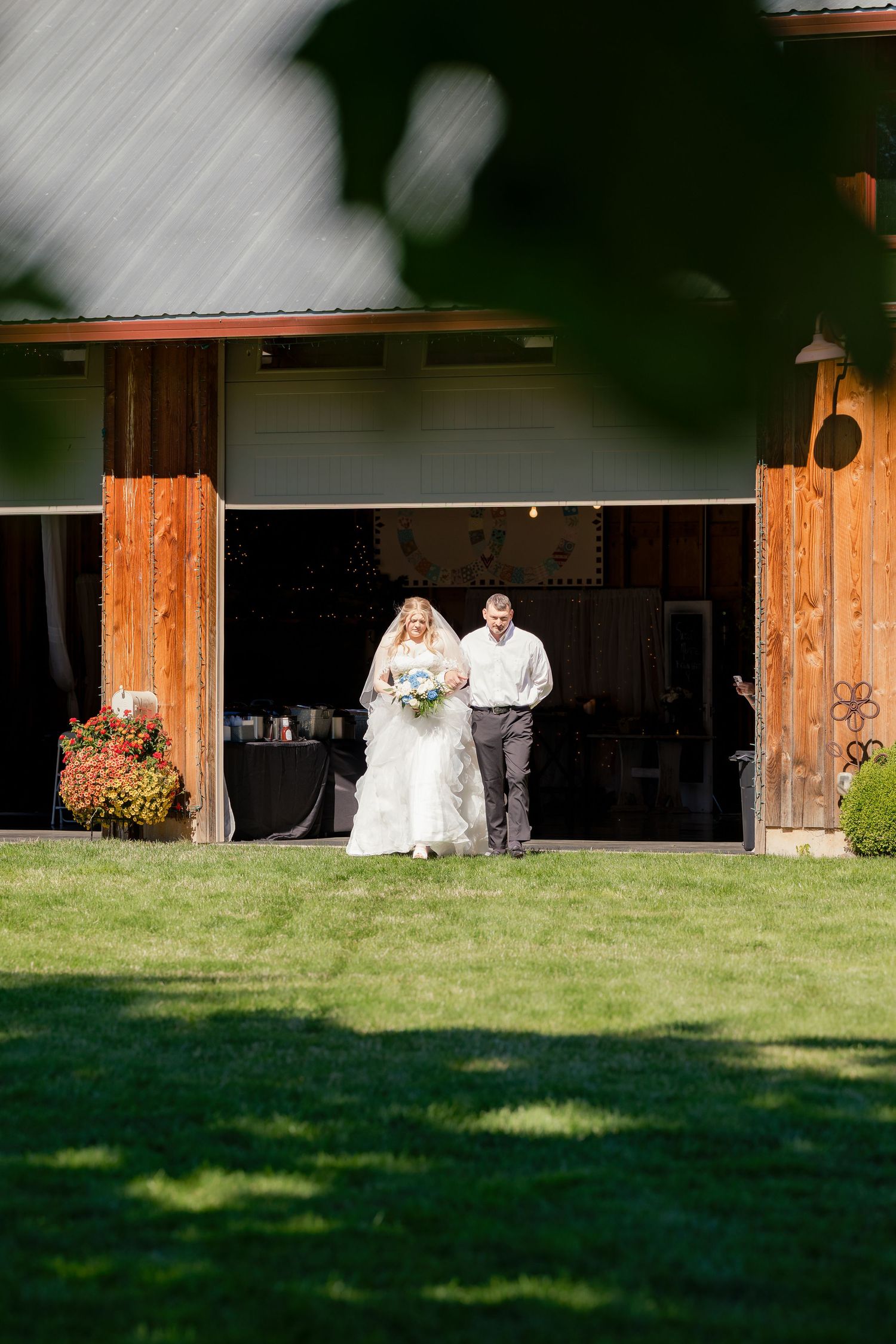 Bride and groom walking out of rustic barn venue onto green lawn during outdoor wedding.
