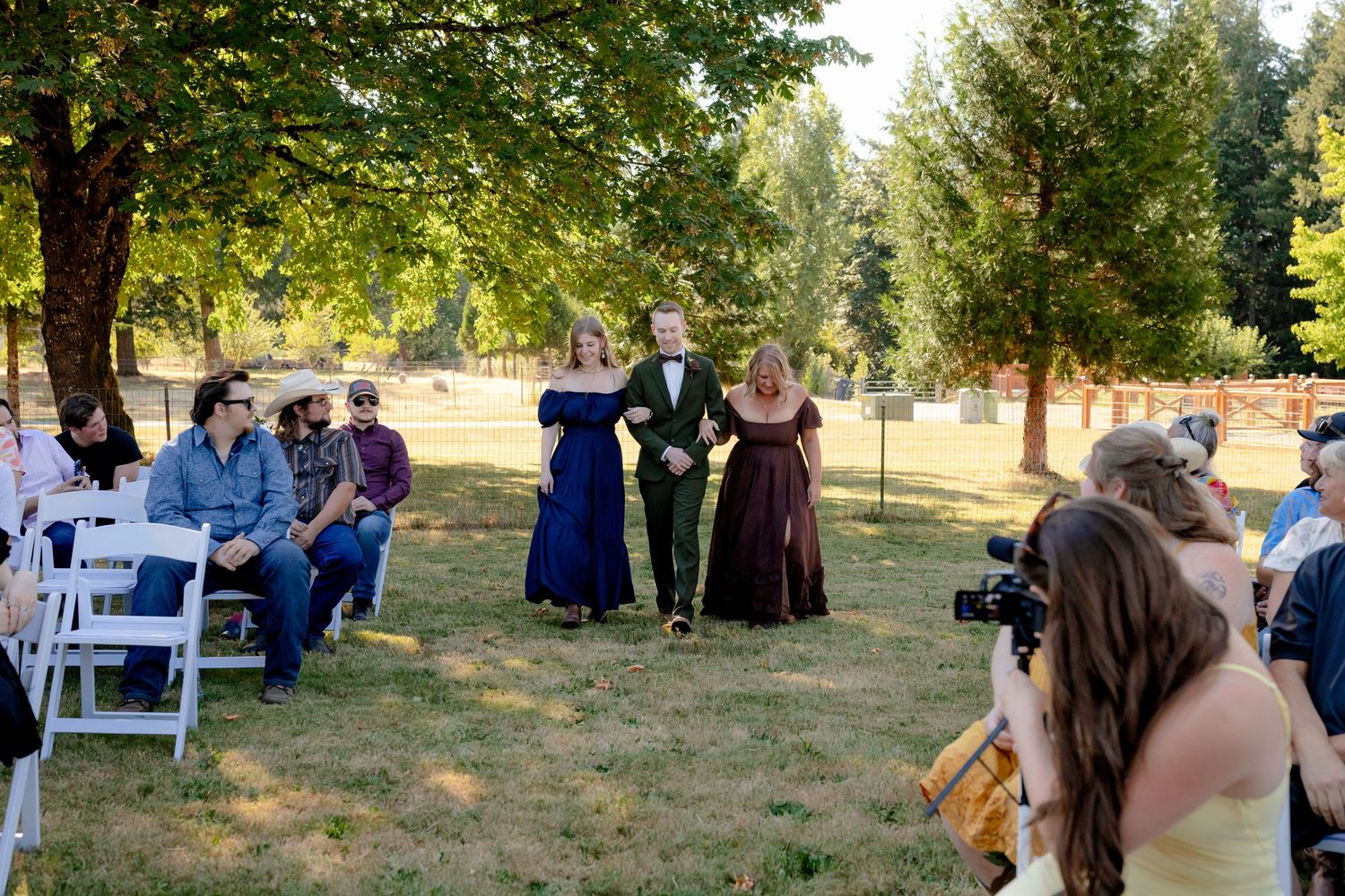 Outdoor wedding ceremony with guests seated on white chairs as the groom walks down the aisle with two attendants.