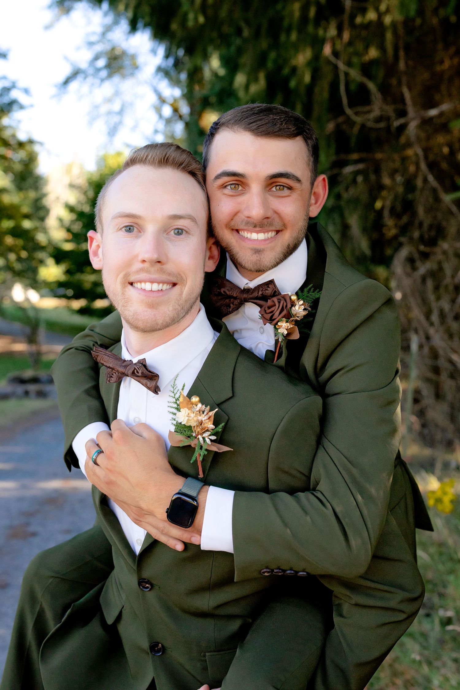 Two grooms in matching green suits smile warmly in a joyful piggyback pose at their wedding.