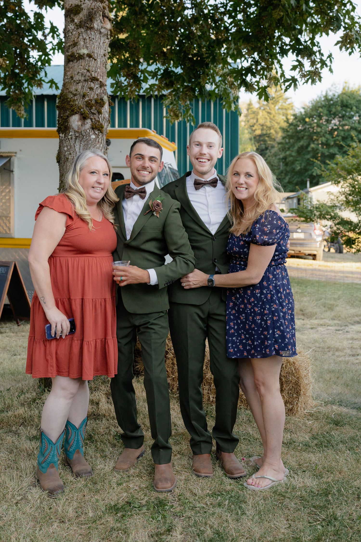 Two grooms in olive green suits pose with two women in colorful dresses at an outdoor rustic wedding.