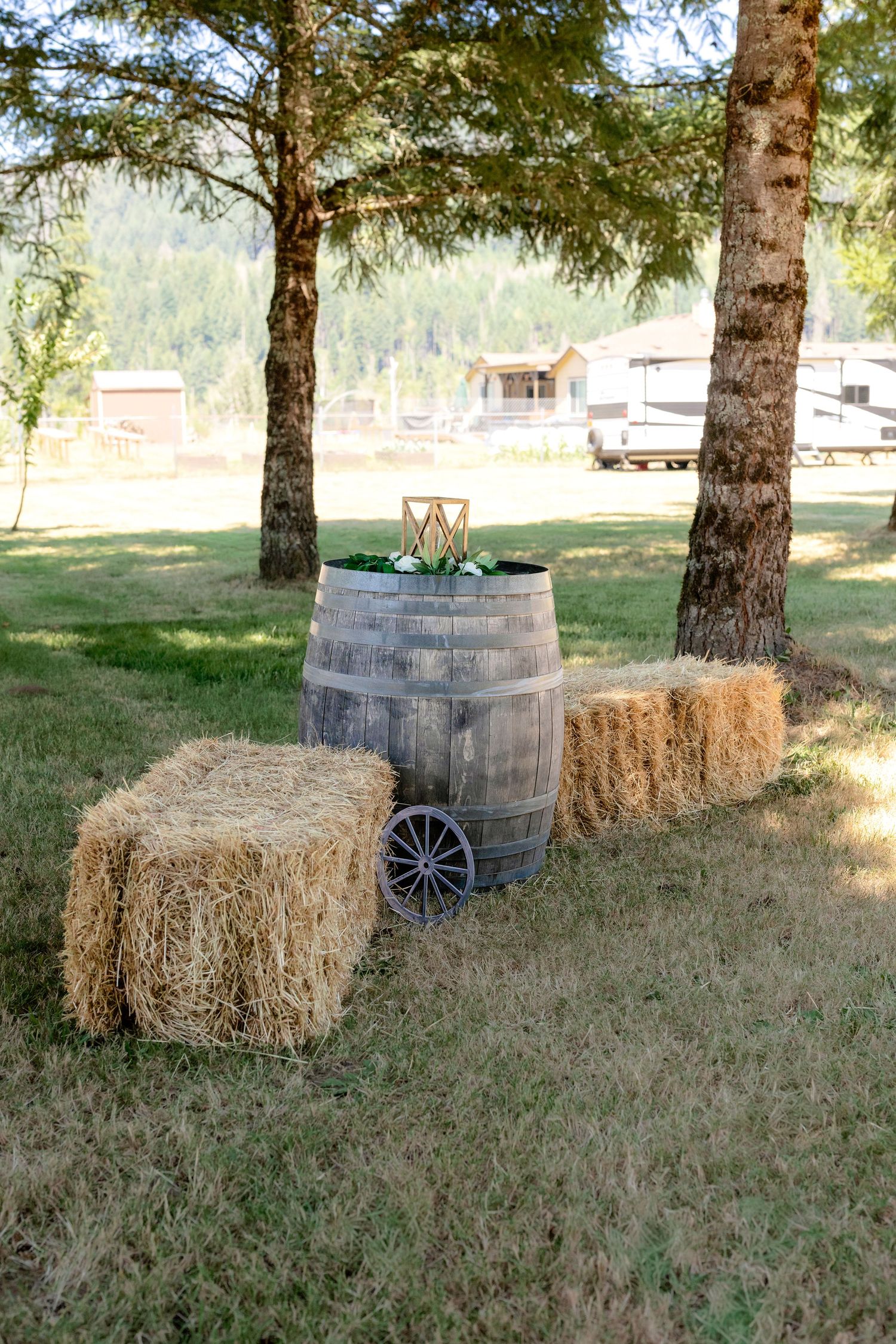 Rustic wedding decor with hay bales and wooden barrel centerpiece under trees at outdoor venue.