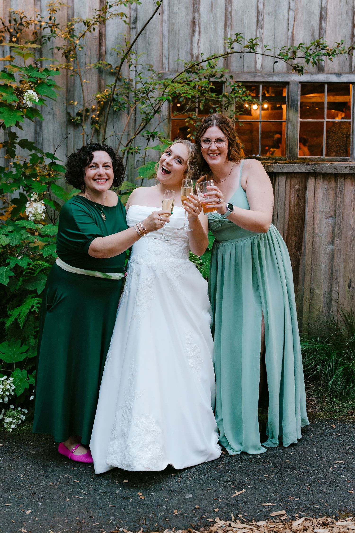Bride in white lace gown toasting champagne with two women in green dresses at outdoor wedding.