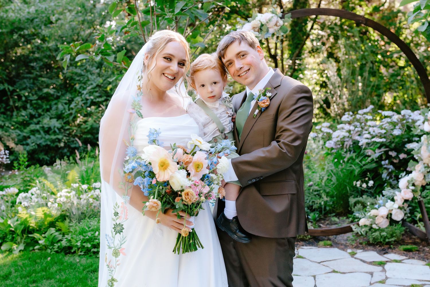 Bride and groom holding baby in garden wedding ceremony with floral bouquet and blooming roses backdrop.