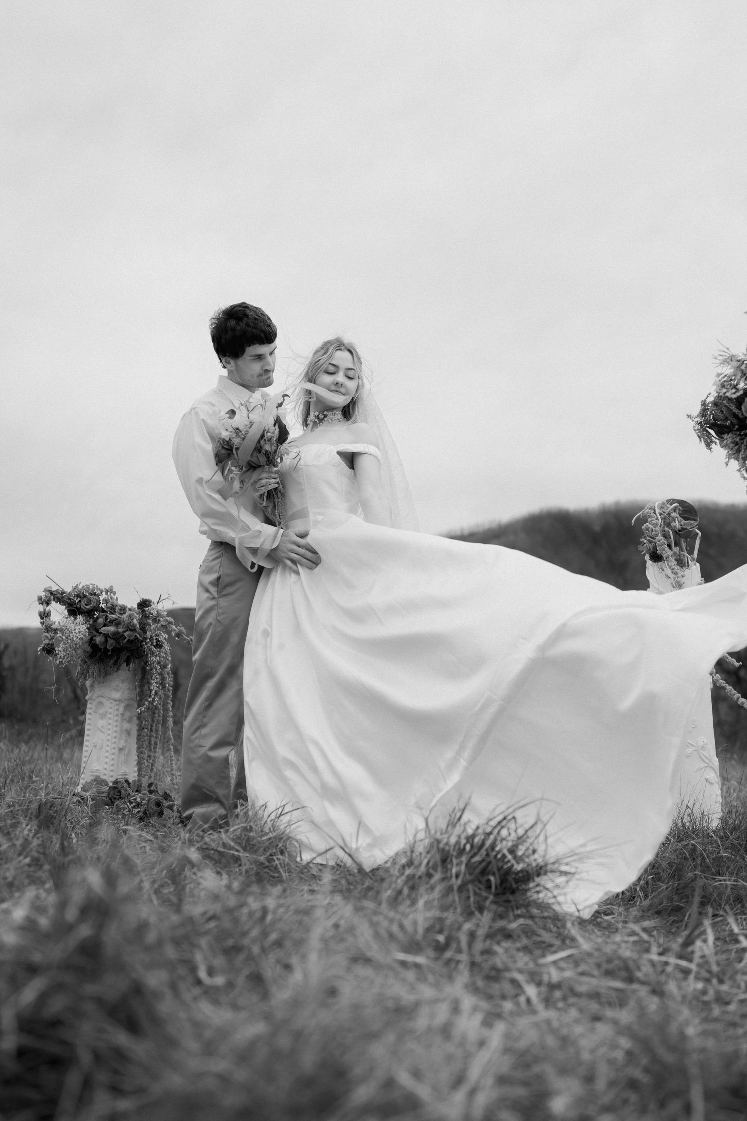 Black and white photo of a couple dancing together in tall grass with flowing white dress.