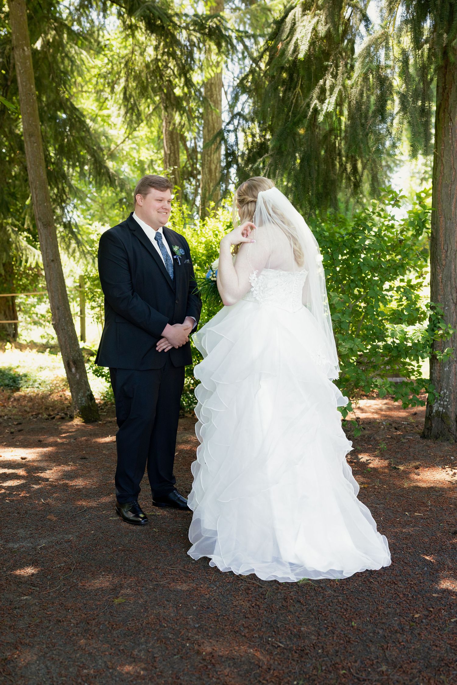 Groom in black suit smiling at bride in layered white gown during first look moment in lush green forest setting.