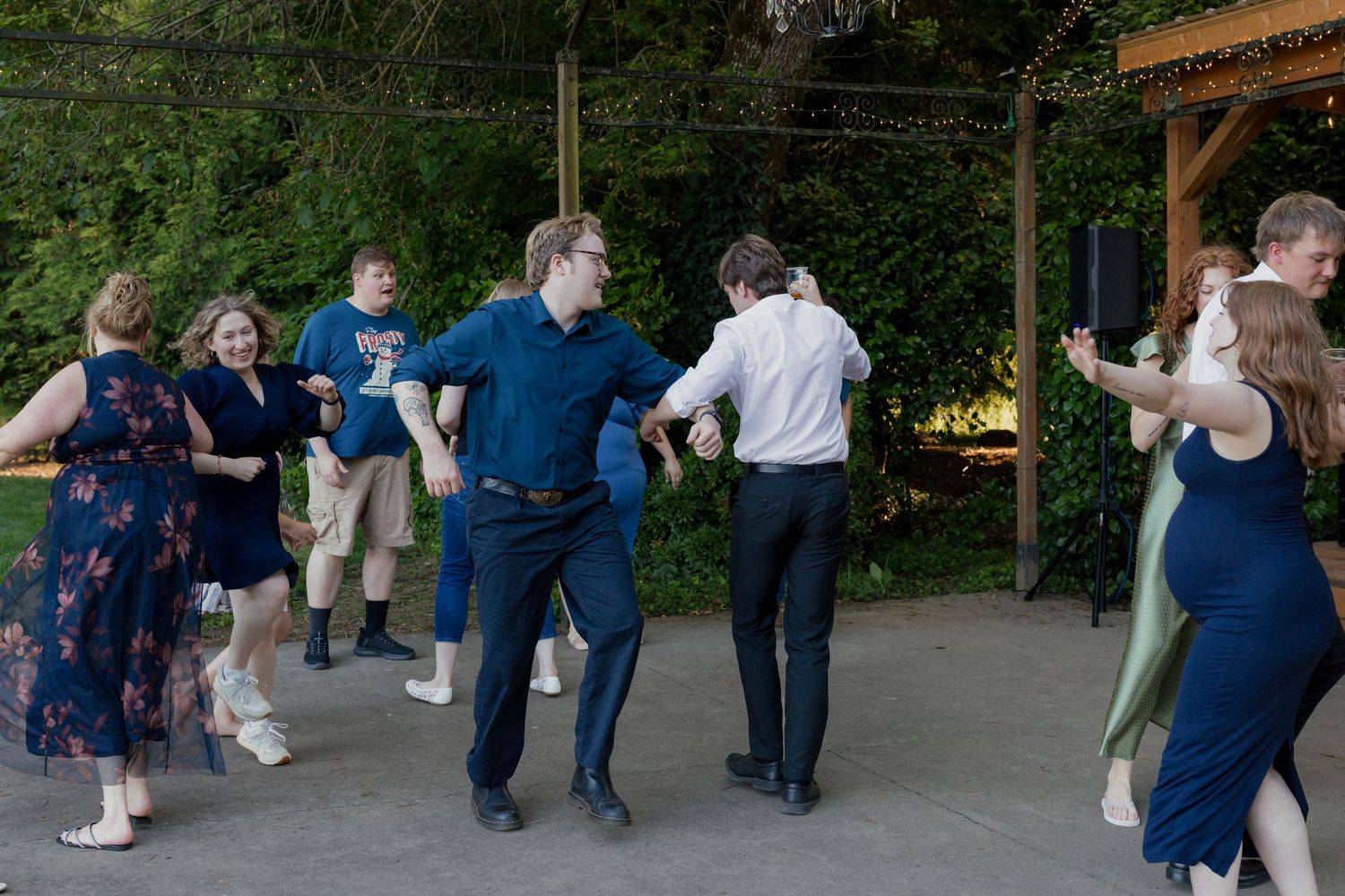 Wedding reception guests dance energetically on outdoor patio at night with DJ speaker visible in background.