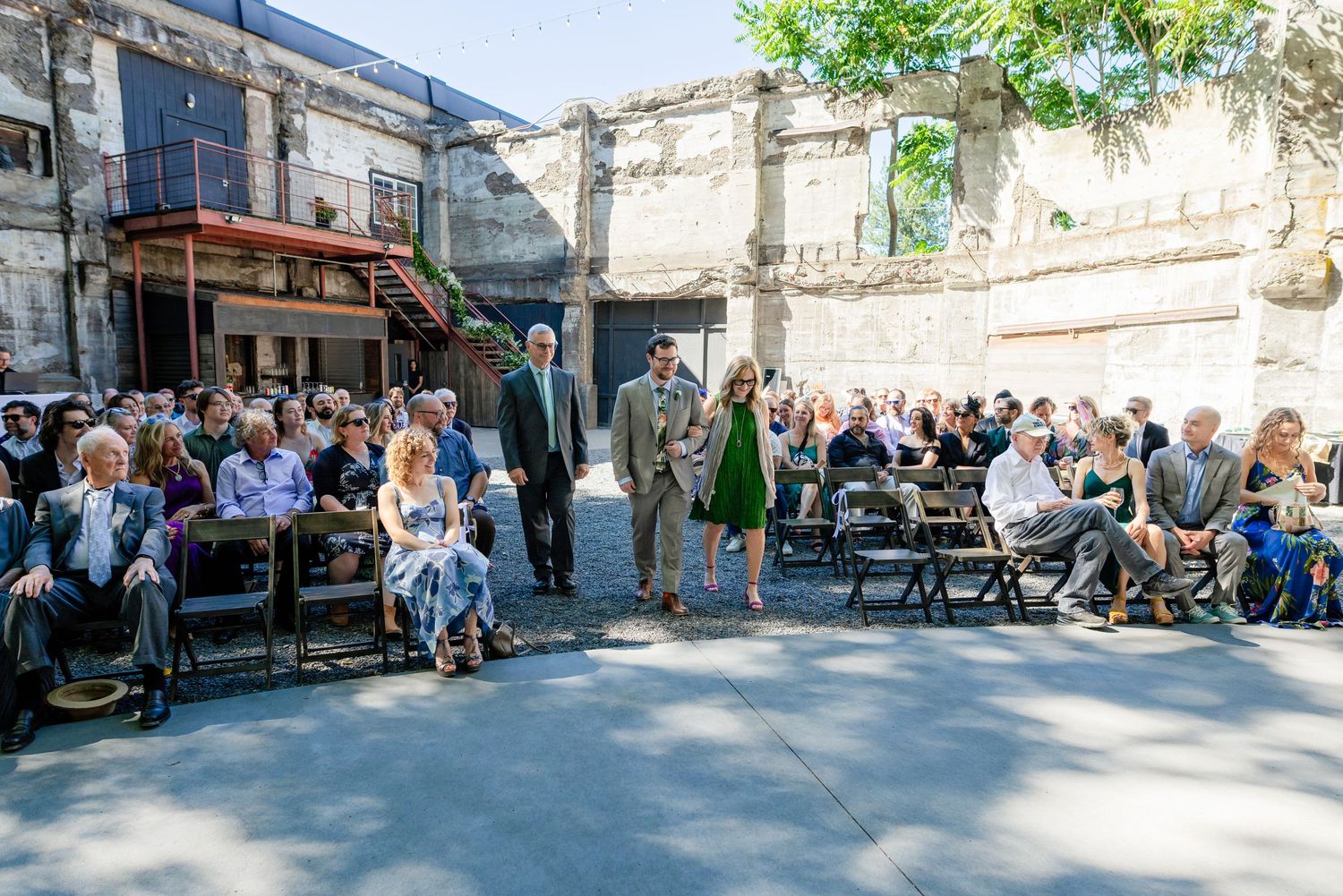 Wedding guests seated outdoors in a rustic urban courtyard venue surrounded by weathered stone walls and trees.