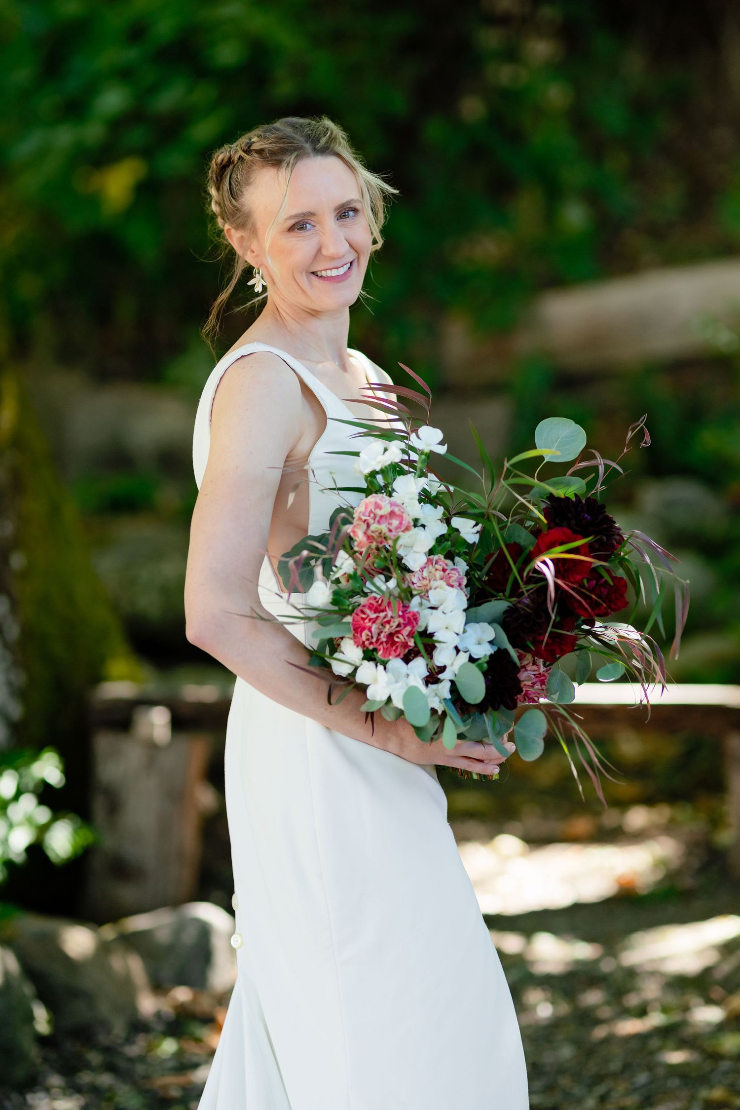 Smiling bride in white gown holds colorful bouquet of pink and red flowers outdoors.