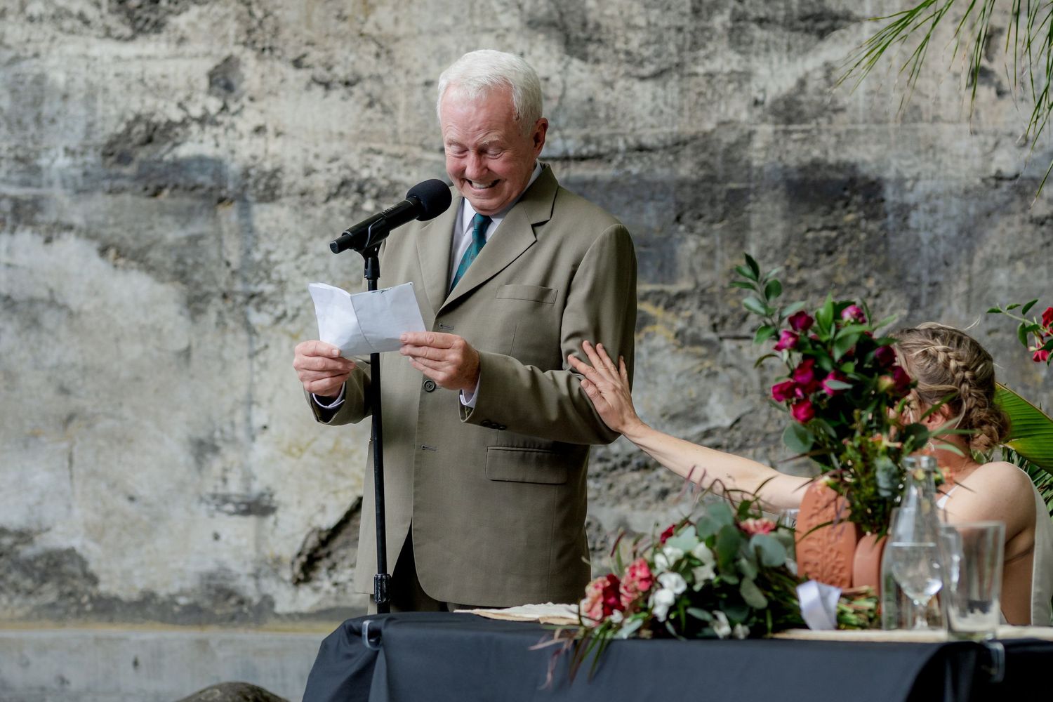 Elderly man in gray suit delivers heartfelt speech at wedding reception microphone.