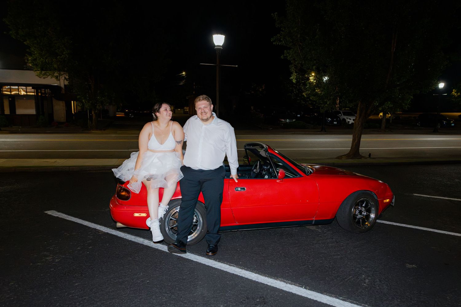 Couple in wedding attire posing beside a red convertible sports car in a parking lot at night.
