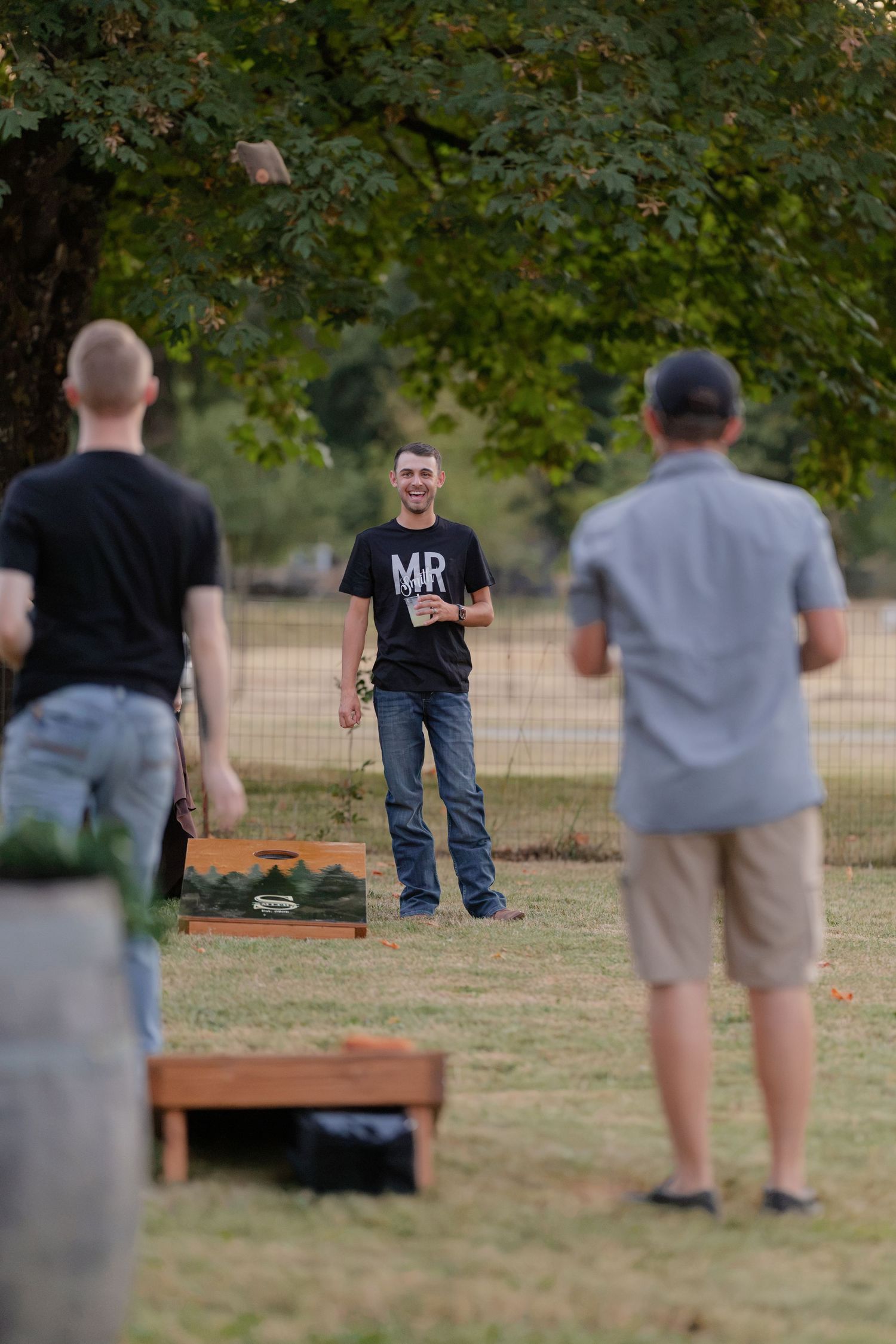 Man in MR t-shirt smiling while playing cornhole at an outdoor wedding reception between two other guests on a grassy lawn.